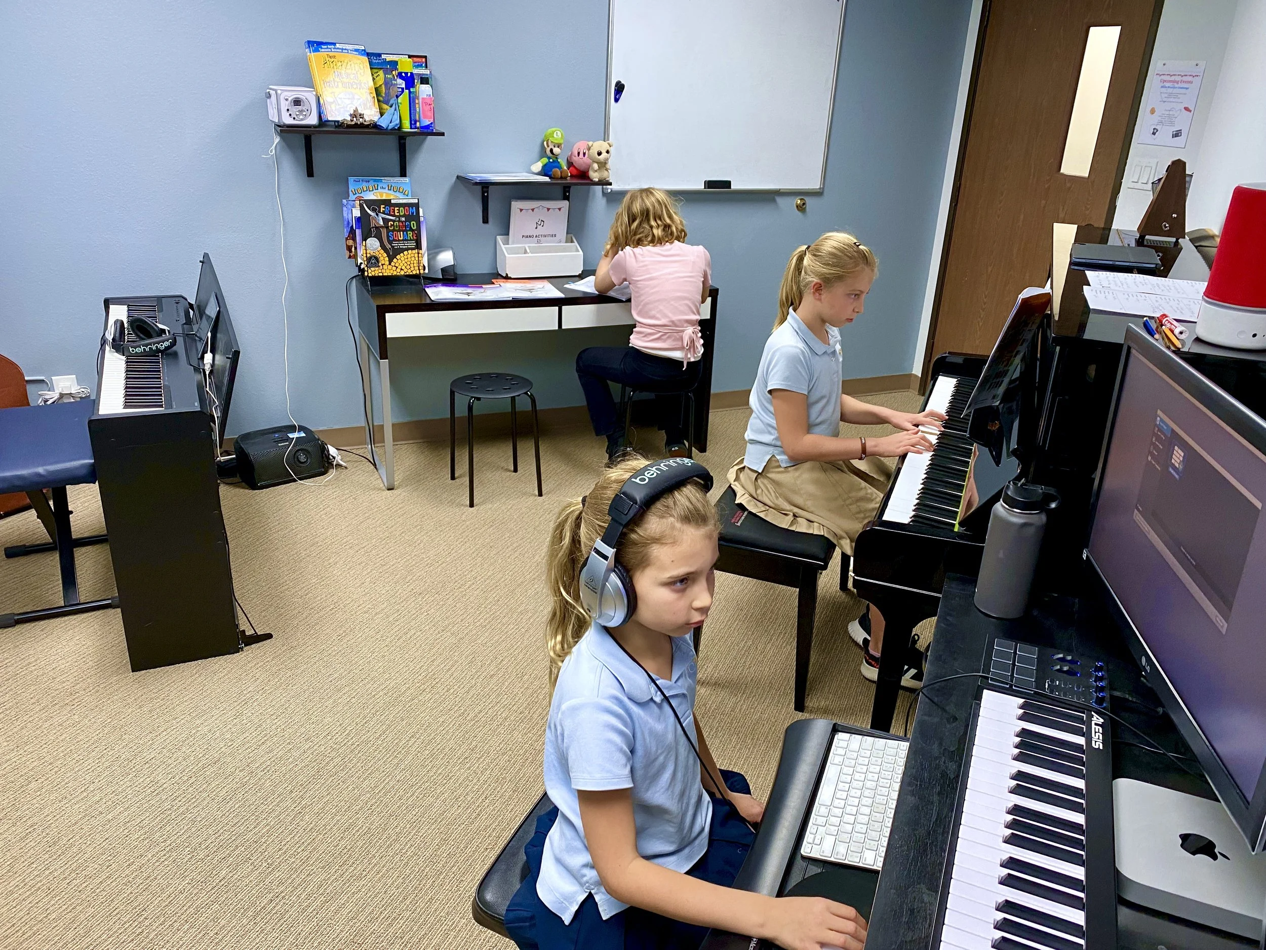 Three young girls are practicing piano in a music classroom. One with headphones sitting at a keyboard in the foreground, and two others playing on pianos in the background.