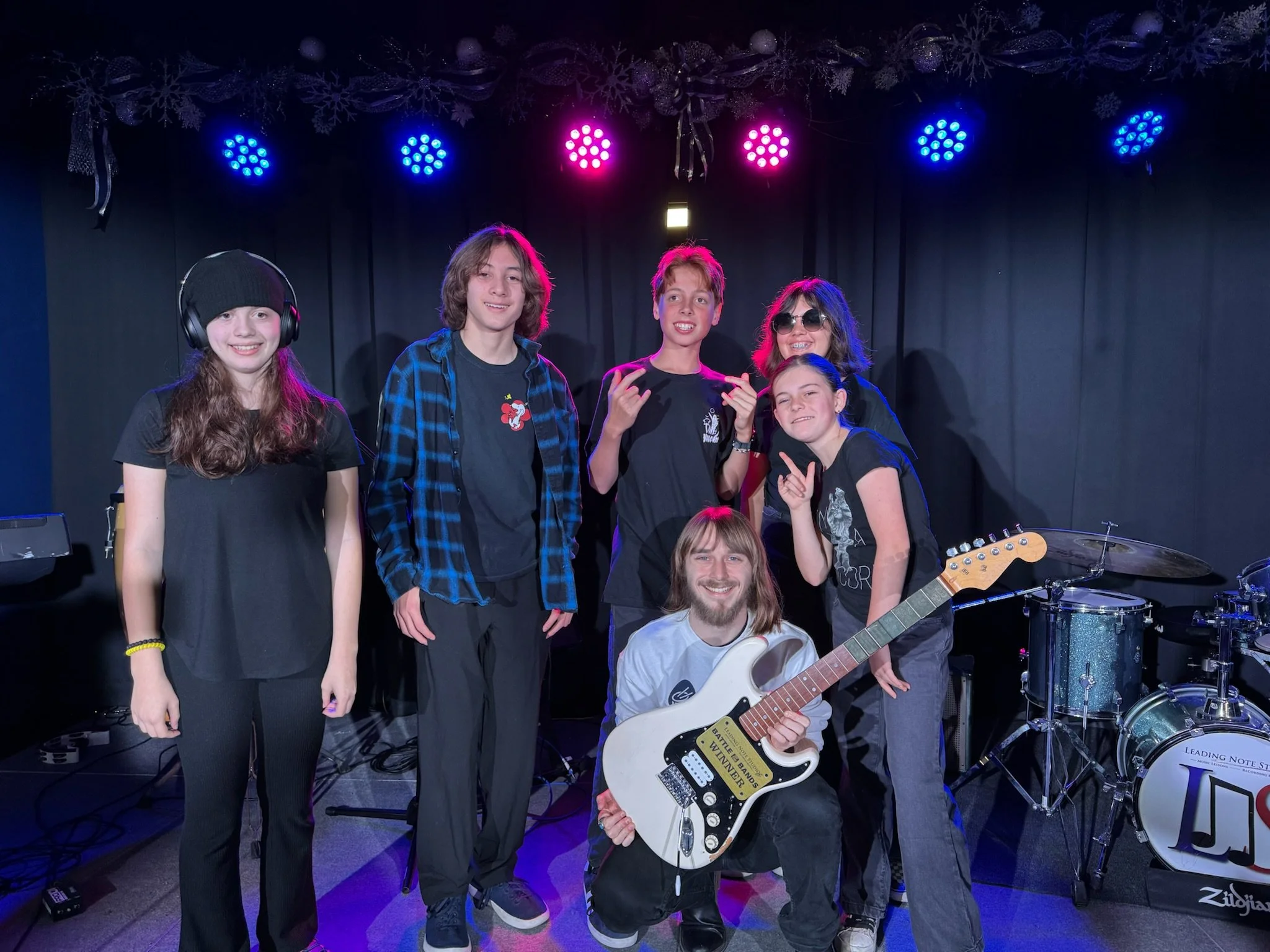 A group of six young people, five standing and one kneeling, posing with musical instruments and over a drum set on a stage with colorful lights.
