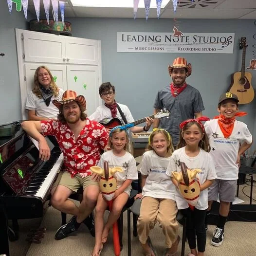 Group of children and adults at a music studio, some wearing cowboy hats and bandanas, with a piano, guitar, and sign in the background.