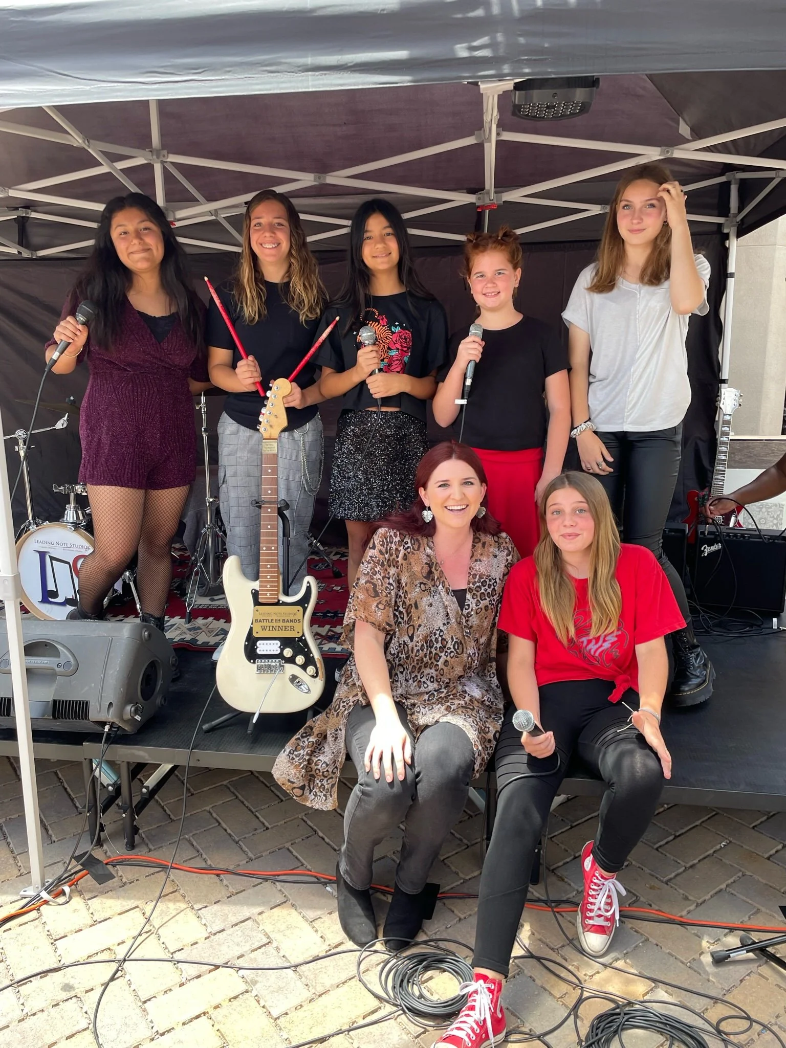 Group of young girls and women on stage with musical instruments and microphones, posing for a photo at an outdoor event.