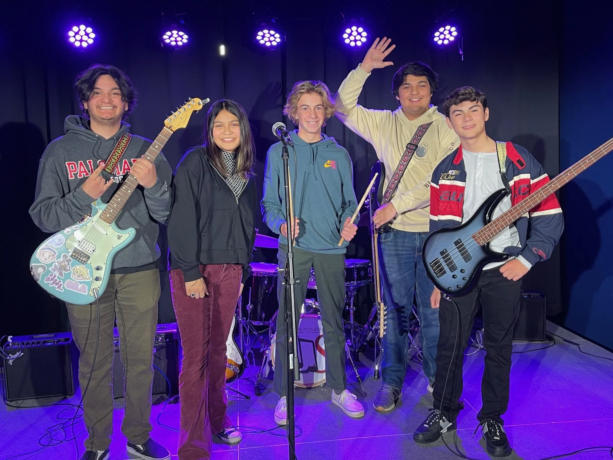 A band of five young people on stage with musical instruments, smiling, under purple stage lights, with drums and equipment in the background.