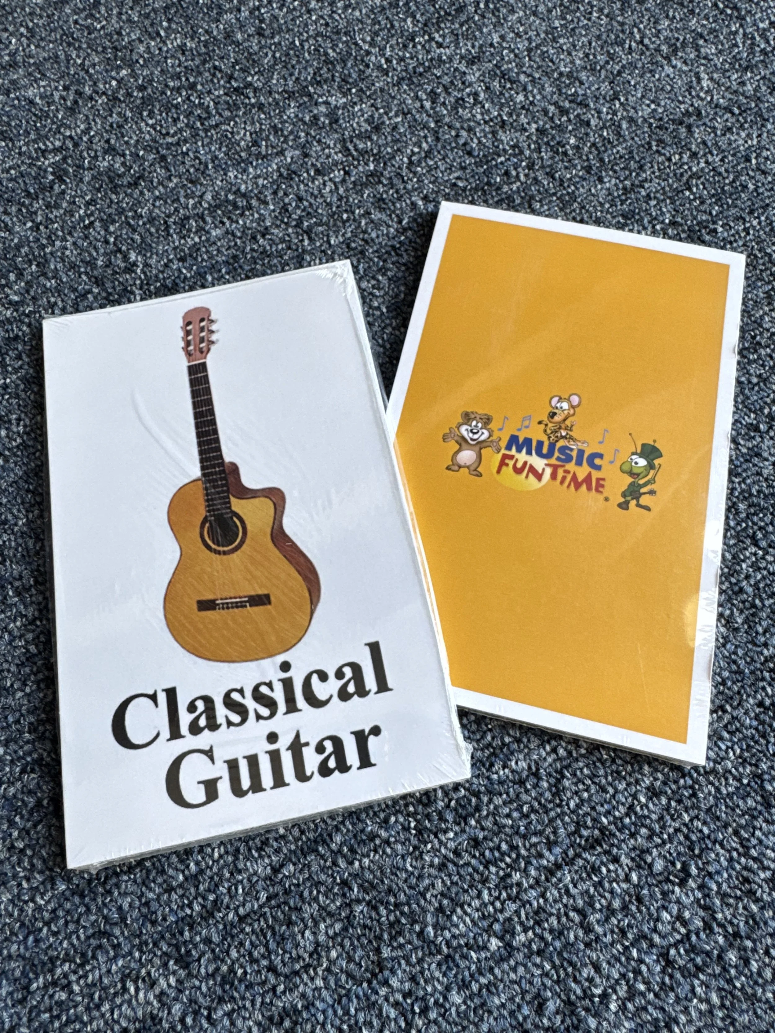 Two educational flashcards on a gray carpet. One card shows a wooden classical guitar with the text 'Classical Guitar', and the other card features colorful cartoon characters with the text 'Music Fun Time'.