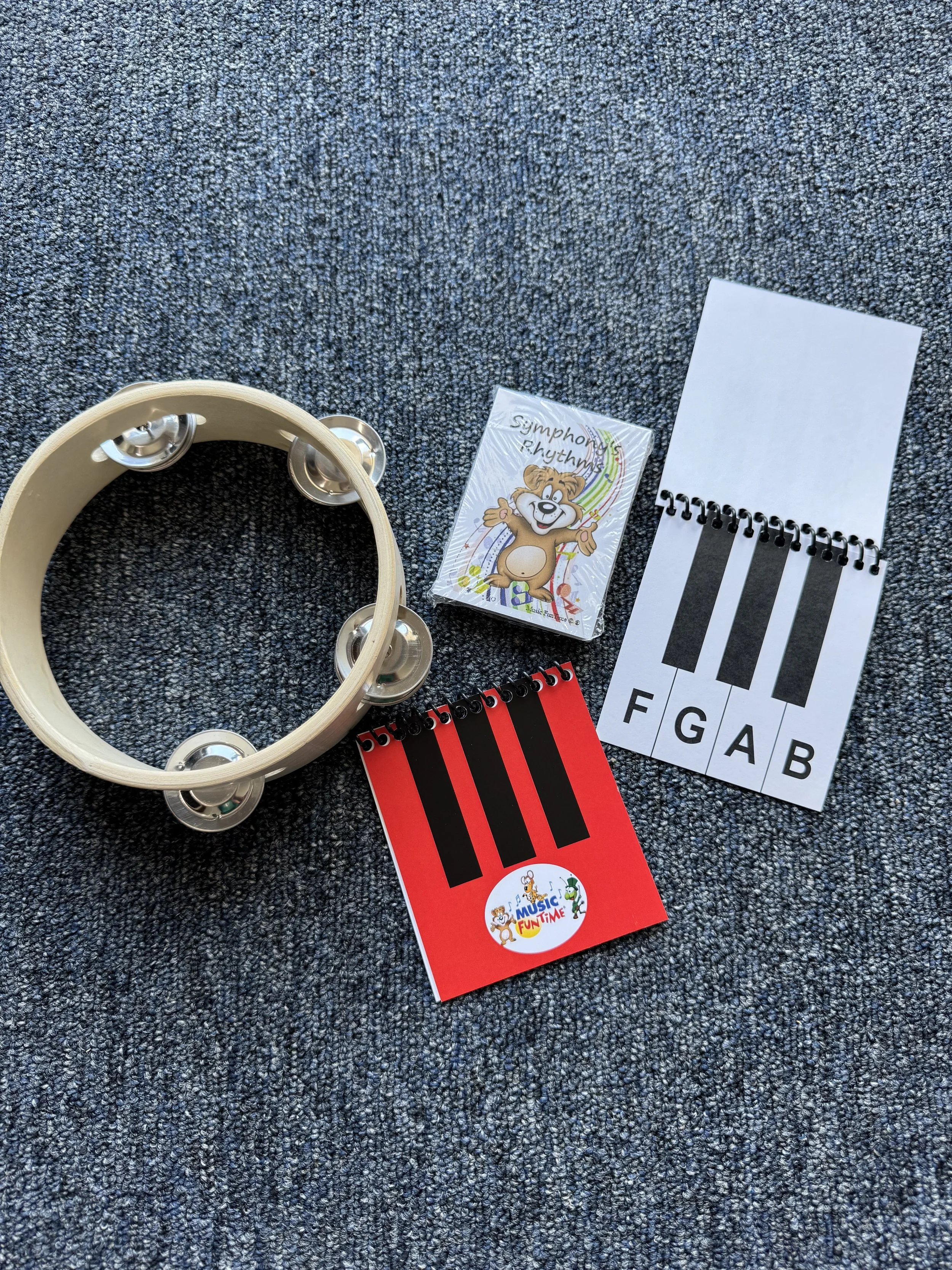 A tambourine, two miniature piano key cards labeled F, G, A, B in black and white, and colored with black stripes, and a small packet with a cartoon bear and the words 'Symphony Rhythms' are arranged on a gray carpeted surface.