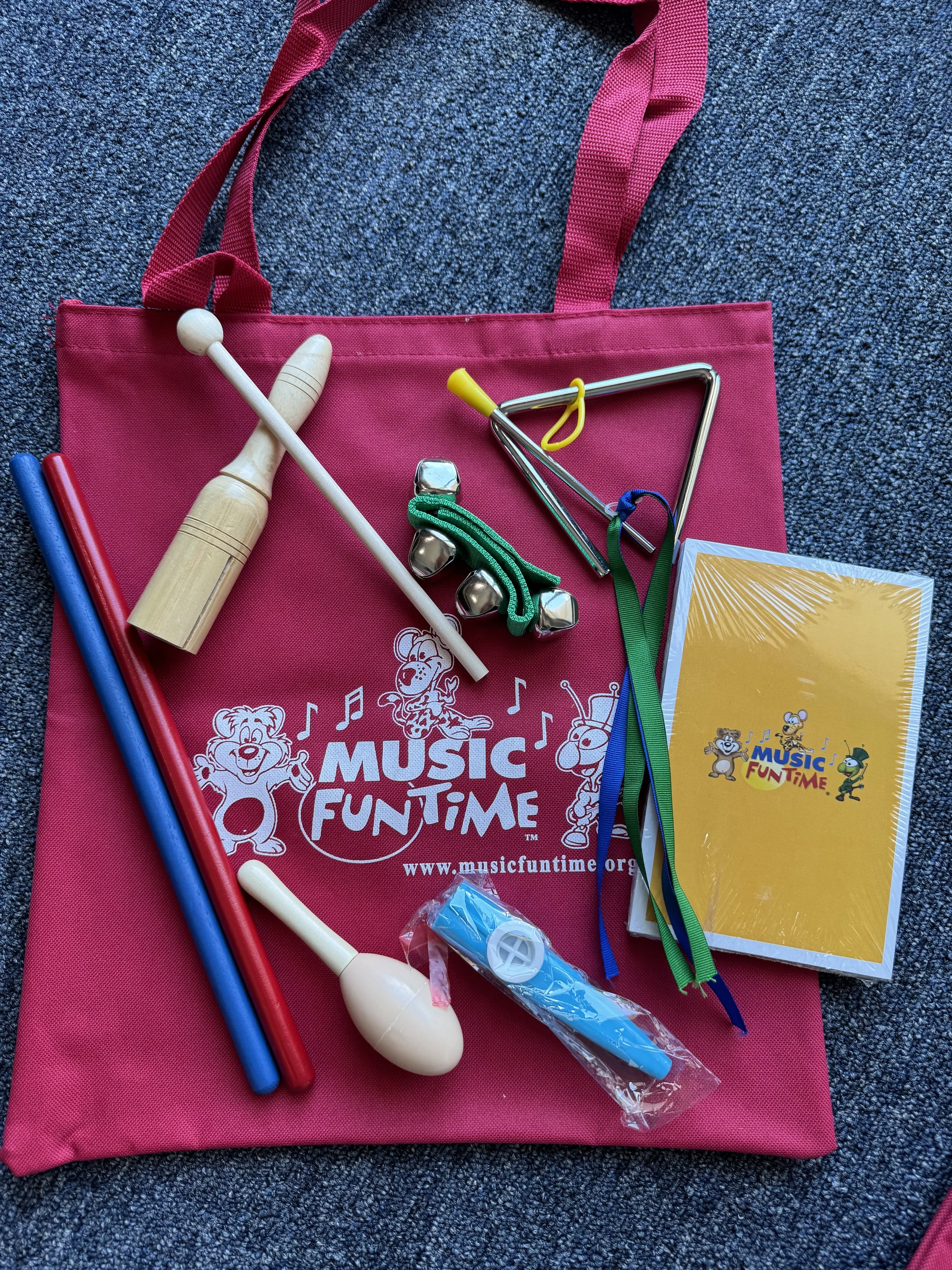 A pink Music Fun Time tote bag on a gray carpet, with various percussion instruments inside including two drumsticks, a tambourine, a small blue bat, a white maraca, metal jingle bells, and a triangle. There is also a yellow book with the Music Fun T