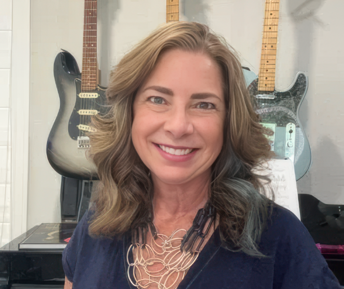 A woman with shoulder-length wavy brown hair smiling at the camera, with electric guitars hanging on the wall behind her.