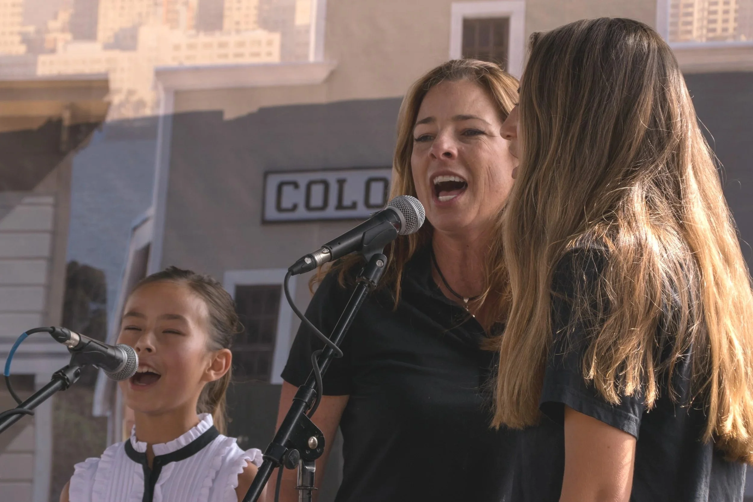 Three women singing into microphones on an outdoor stage with a building in the background.