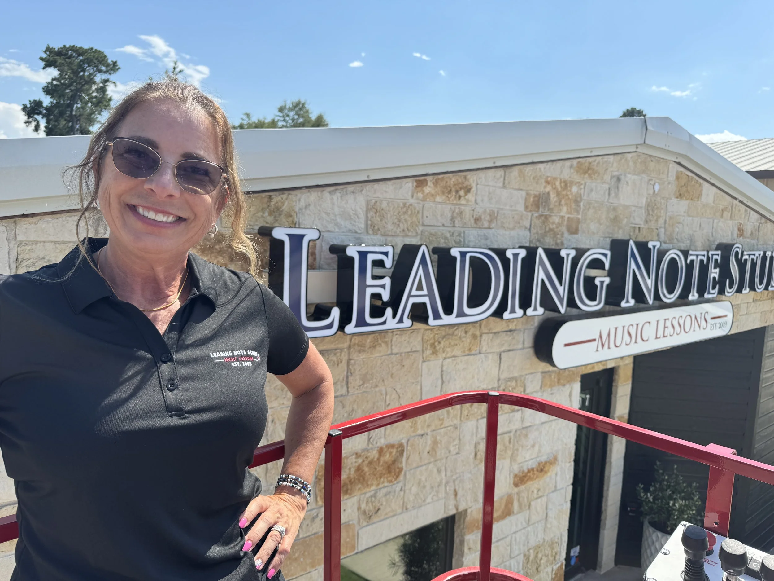 Smiling woman in sunglasses standing outside Leading Note Studio with a sign that reads 'MUSIC LESSONS'.