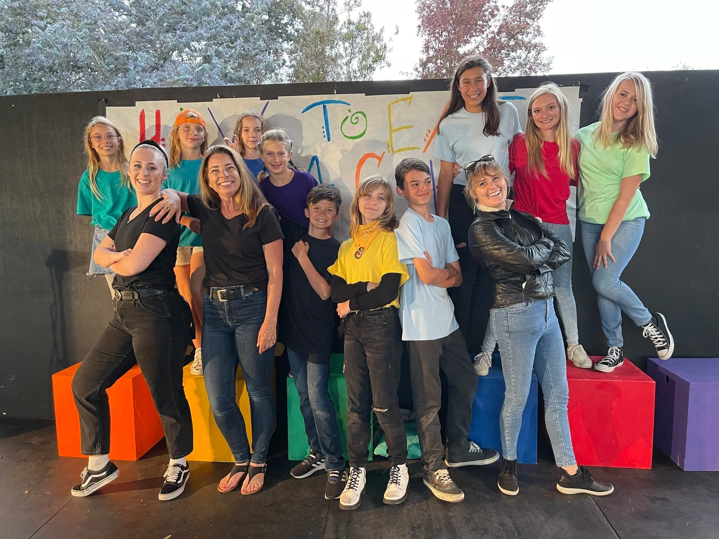 A group of children and two women posing together on a stage, with a colorful backdrop and large colored blocks, during a group event or performance.