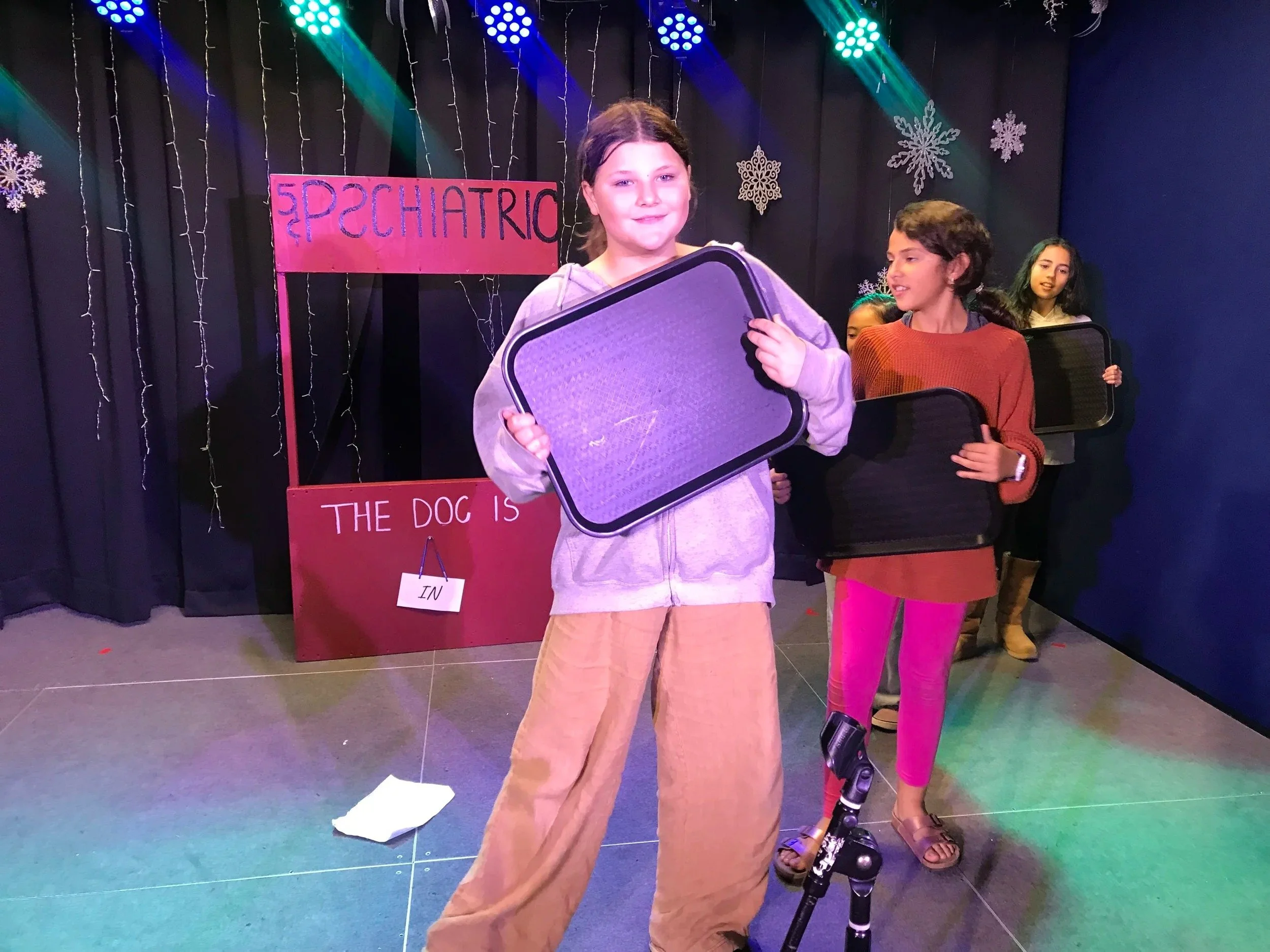 Group of girls standing on a stage decorated with snowflakes, holding clipboards, in front of a pink sign that reads 'Epediatric,' with smaller text underneath. Stage lights above and colorful lighting on the floor.