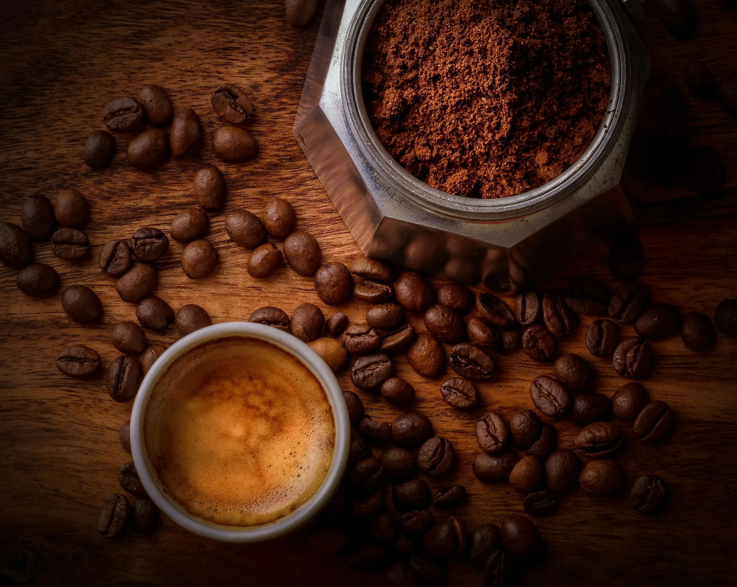 Ground coffee in a container, coffee beans scattered on a wooden surface, and a cup of espresso.
