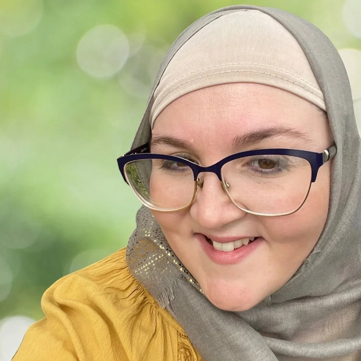 Close-up of a Muslim woman smiling, wearing glasses, a beige hijab, and a yellow top with decorative detailing on the sleeve, against a blurred green background.
