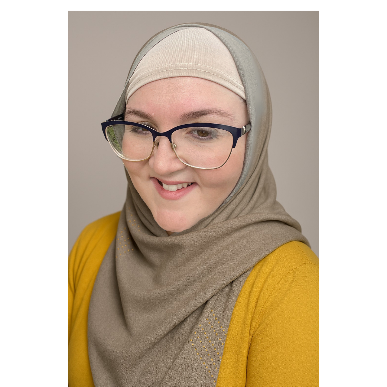 A woman wearing a beige hijab, large glasses, and a yellow outfit, smiling and looking downward against a neutral background.
