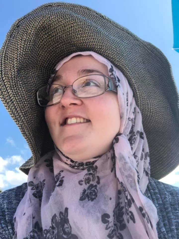 A woman wearing a wide-brimmed straw hat, glasses, and a light-colored scarf with a floral pattern, smiling and looking into the distance against a bright blue sky with a few clouds.