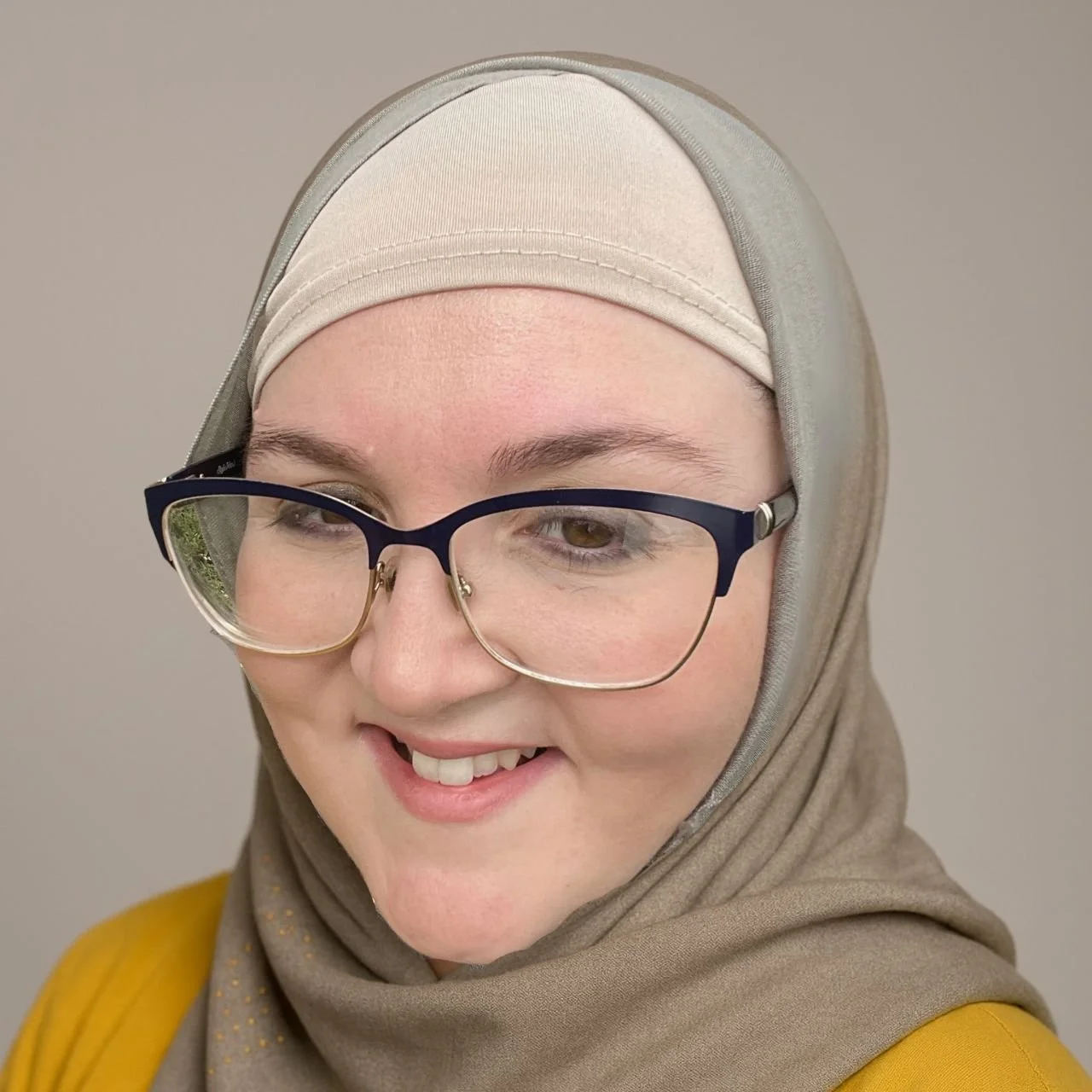 A woman smiling, wearing glasses and a beige hijab, with a beige head covering underneath, and a yellow top.
