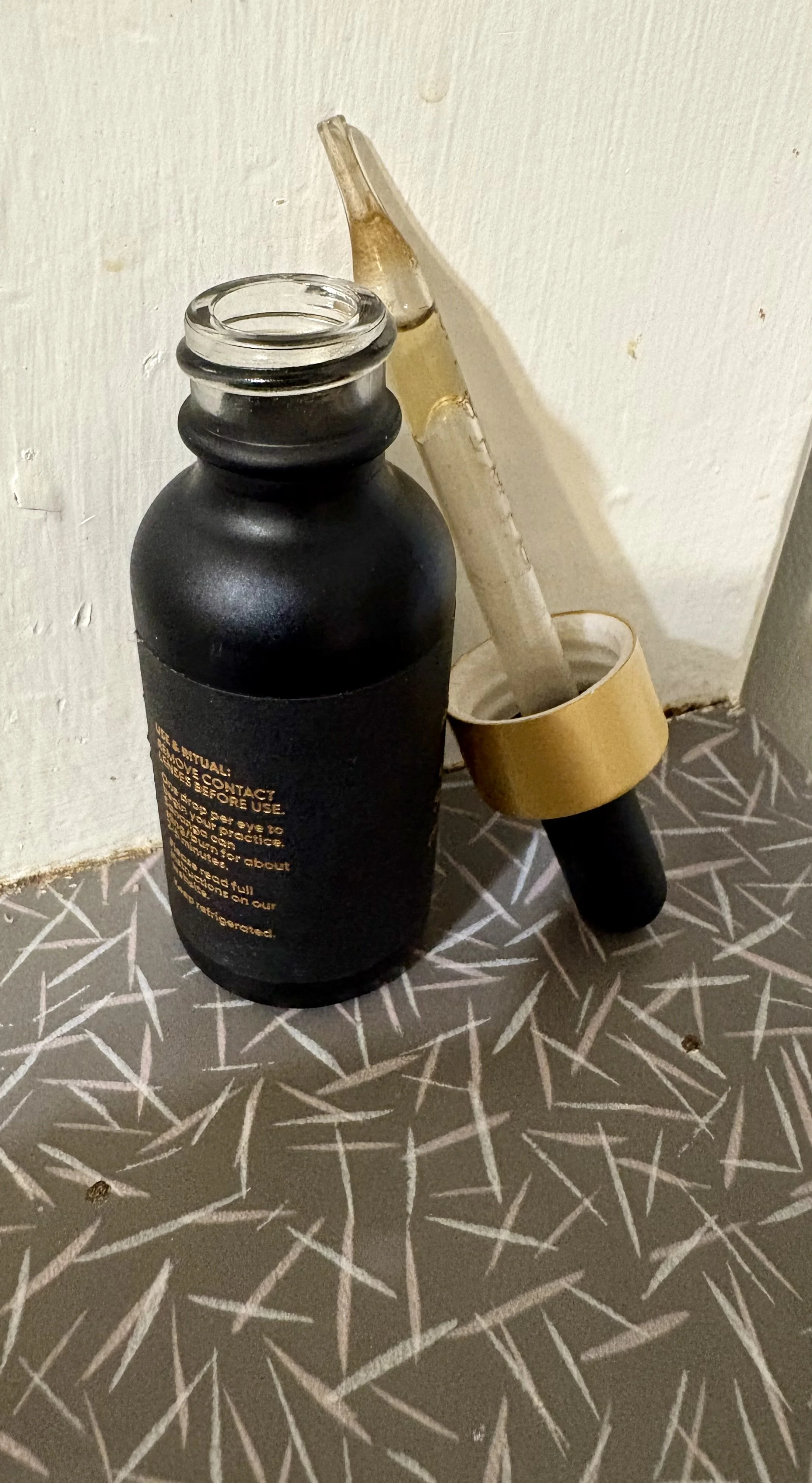 A black dropper bottle with orange instructions on the side, placed on a gray patterned surface near a corner. A glass dropper with a gold cap and a black bulb is resting against the bottle.