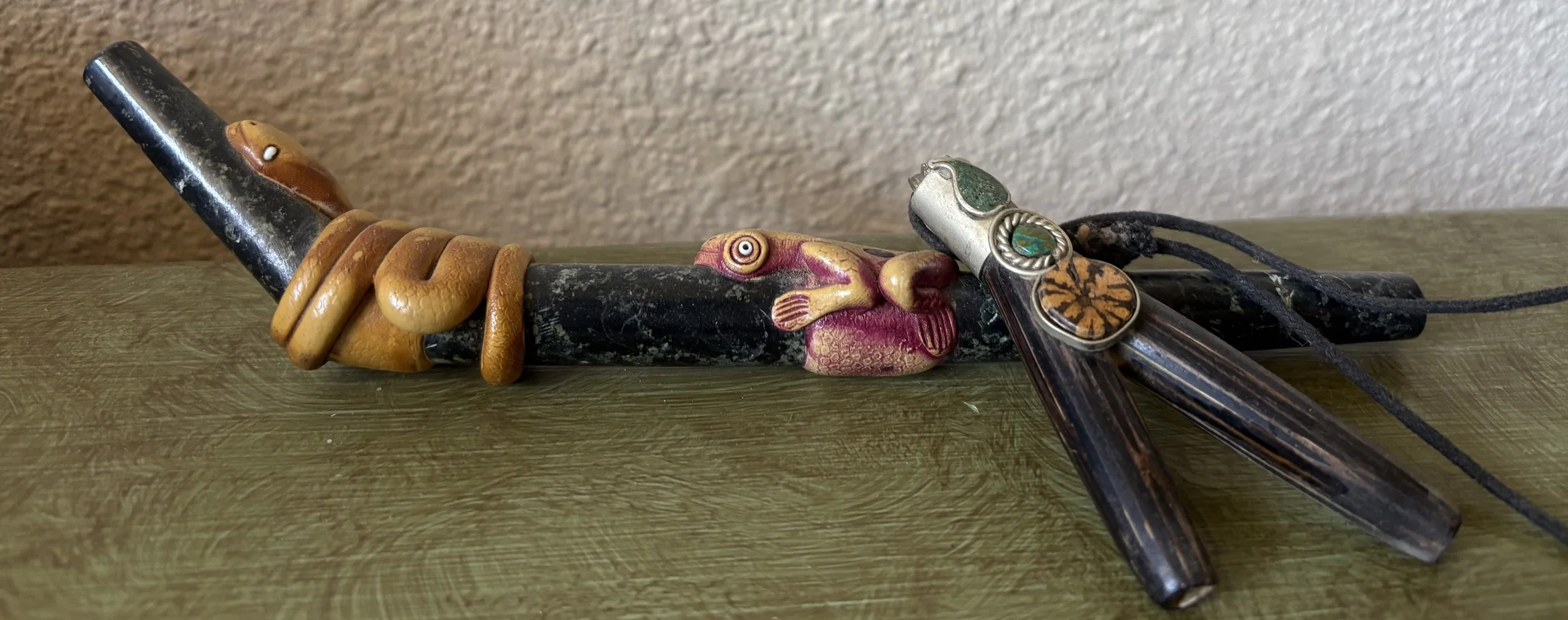 Collection of traditional and decorative items on a green wooden surface including a black pipe with yellow coils, a carved face with a long nose, and several glass and metal jewelry pieces with green and orange accents.