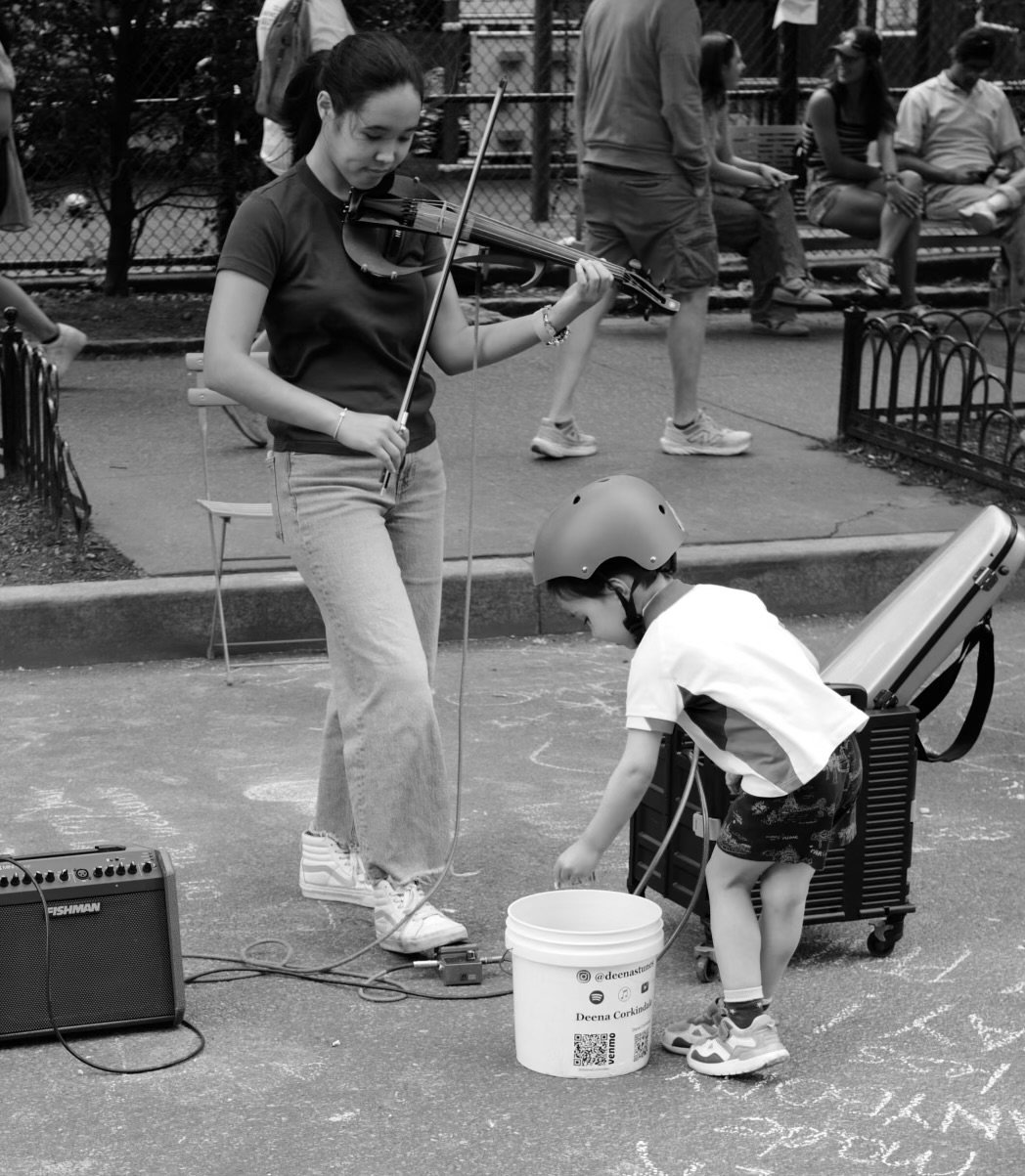 A young girl with a helmet is drawing on the pavement with chalk near a person playing a violin.