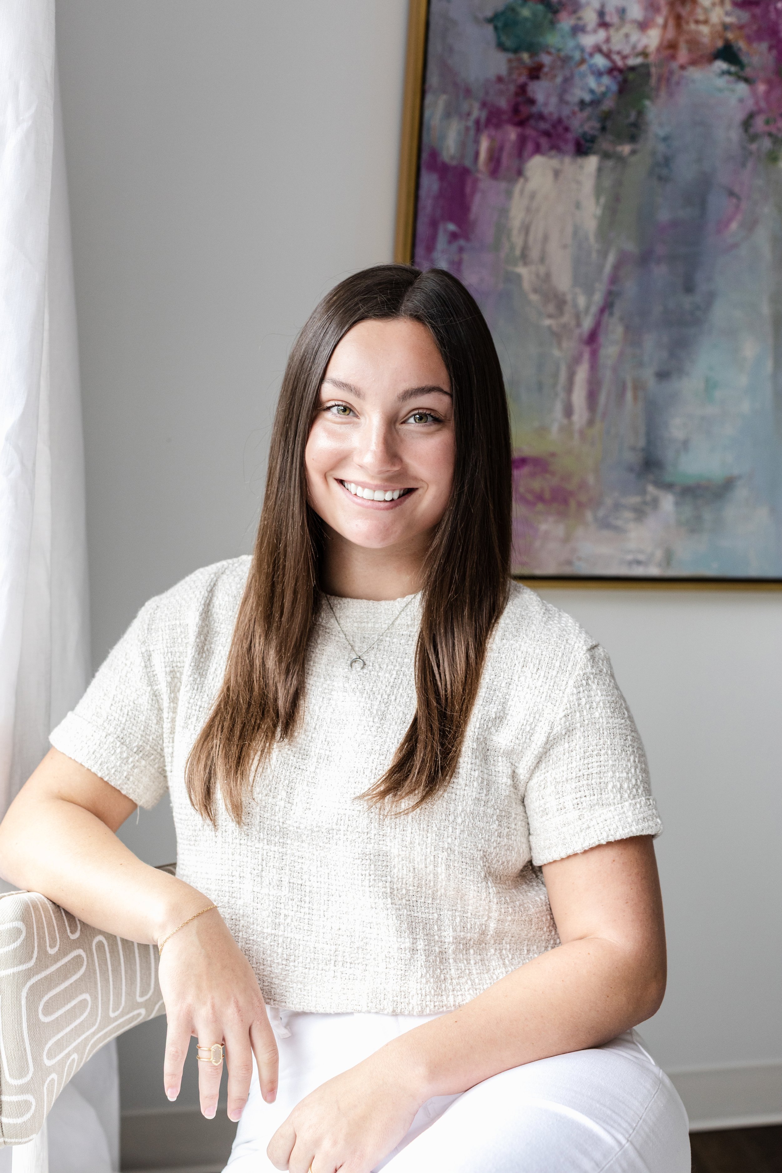 A young woman with long brown hair, smiling, wearing a beige t-shirt, sitting in a room with white walls, a colorful abstract painting on the wall behind her, and a white curtain on the left side of the image.