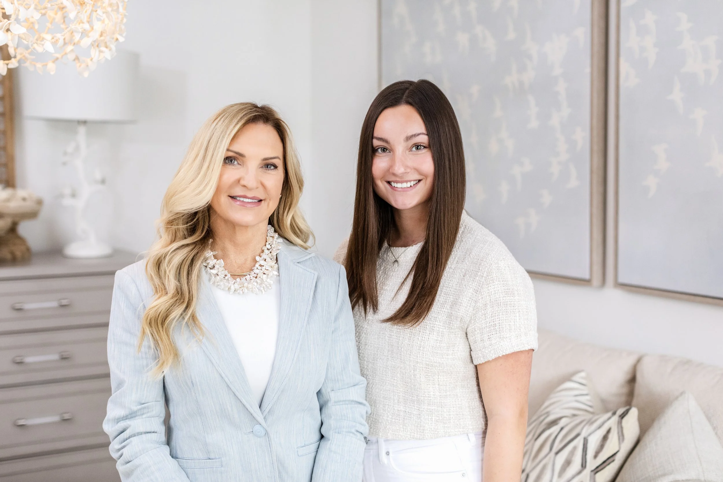 Two women smiling in a well-decorated room with light-colored walls, a patterned pillows on a sofa, and artwork on the wall.