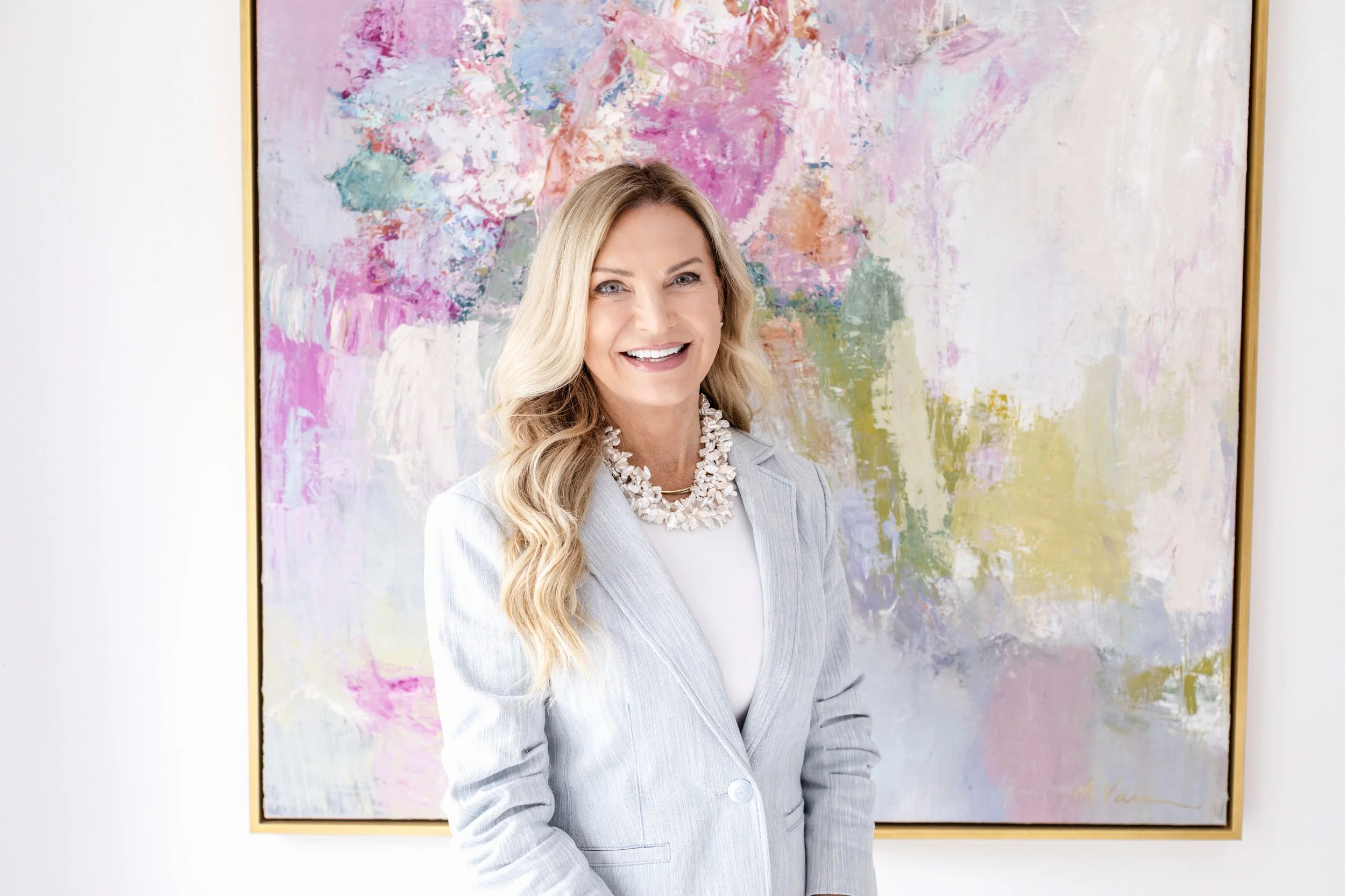 A woman in a light gray blazer and white dress shirt smiling in front of an abstract colorful painting with pink, green, and blue hues.