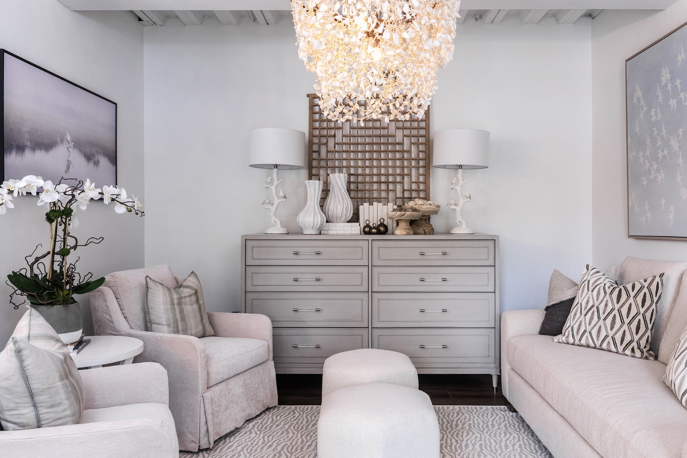Living room with light gray furniture, white decorative lamps, vases, artwork, and a chandelier, with a light-colored rug.