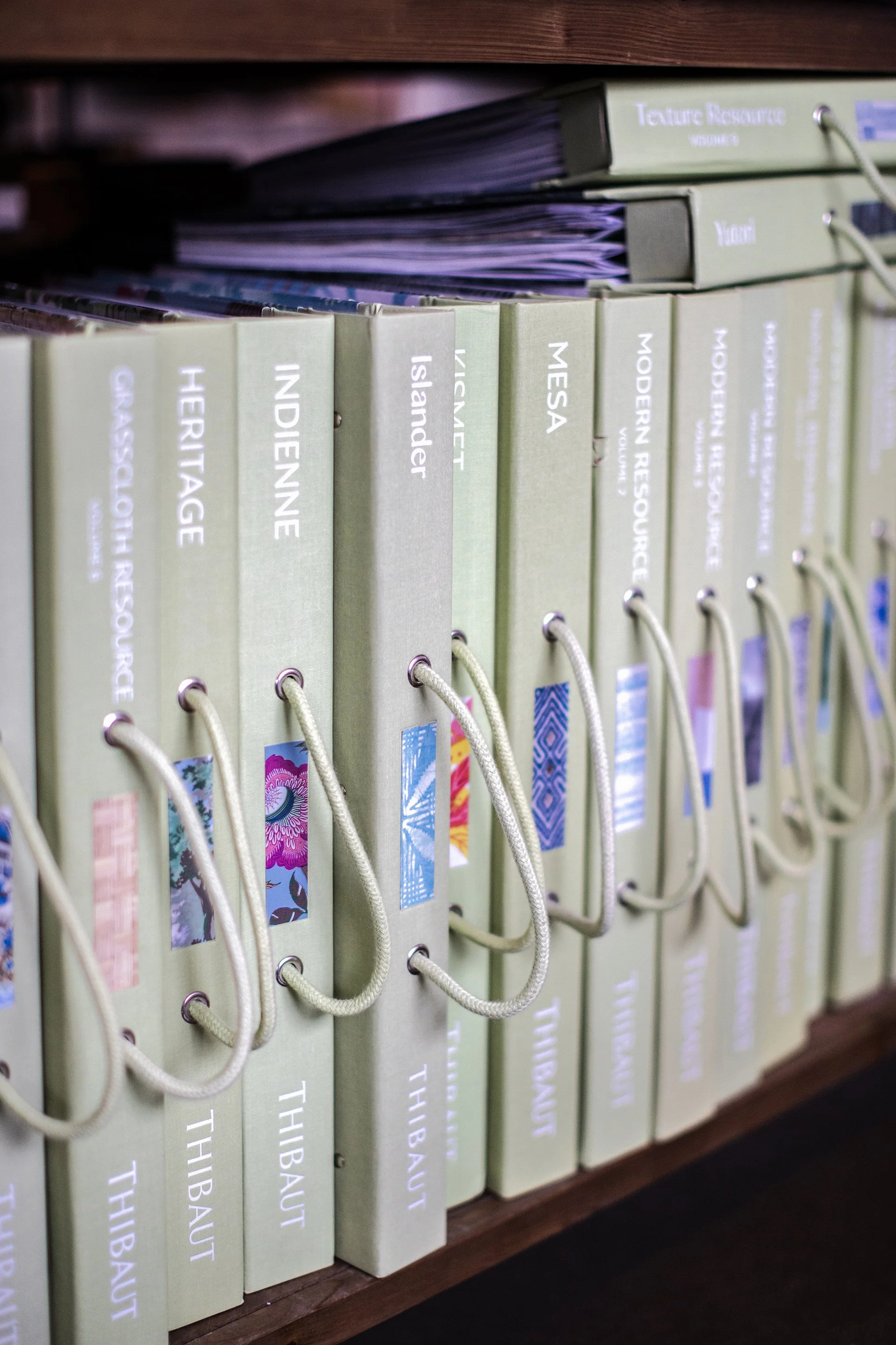 Shelf filled with binders labeled with titles such as 'Heritage,' 'Indienne,' 'Islander,' 'MESA,' and 'Modern Resources,' each with colorful decorative stickers and beige cords attached.