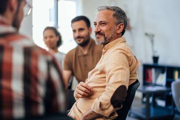 Mature man in group therapy smiling and listening to someone speak.