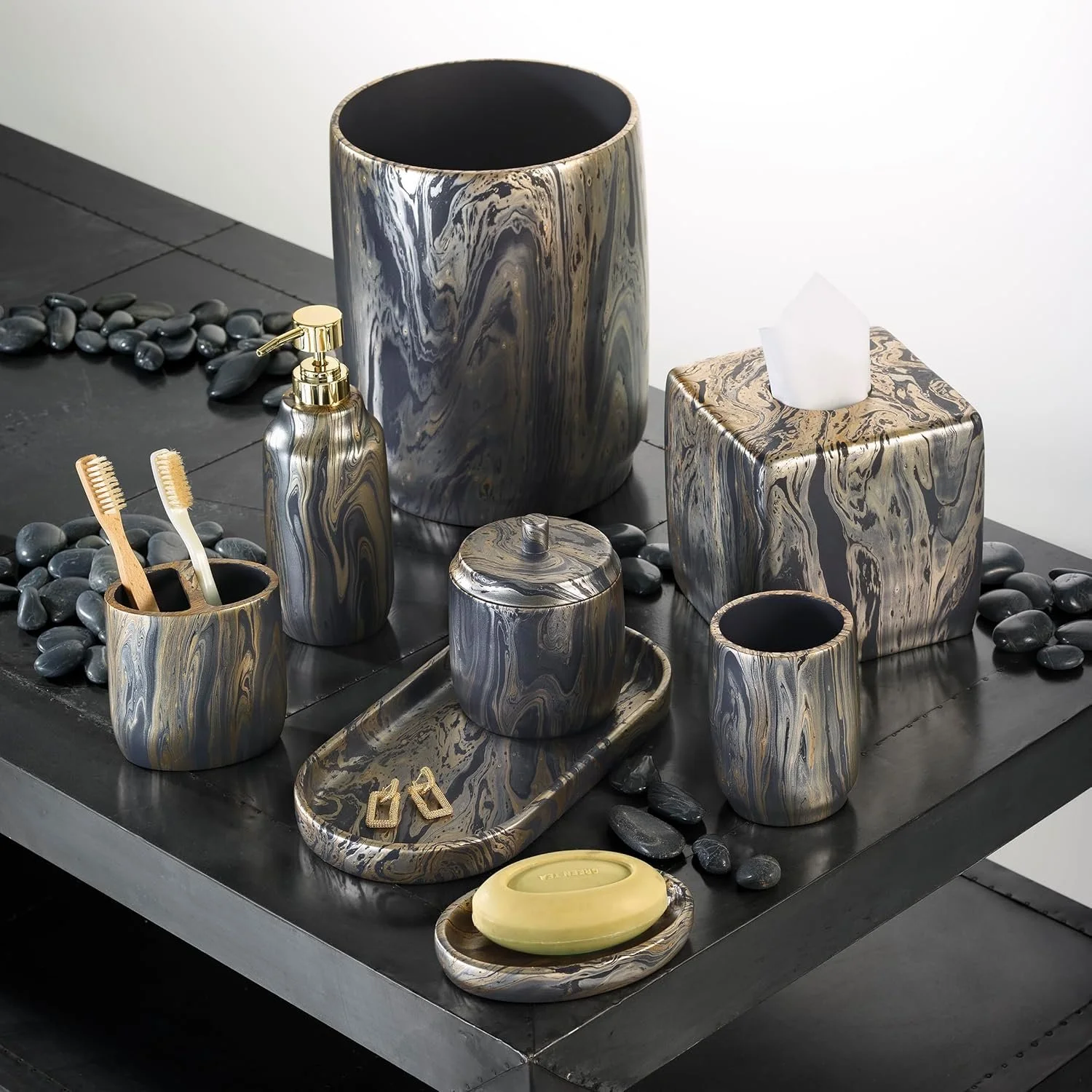Decorative bathroom accessories with a marble pattern, including a soap dispenser, tissue box, small cup, tray, and containers, arranged on a black table with black stones nearby.