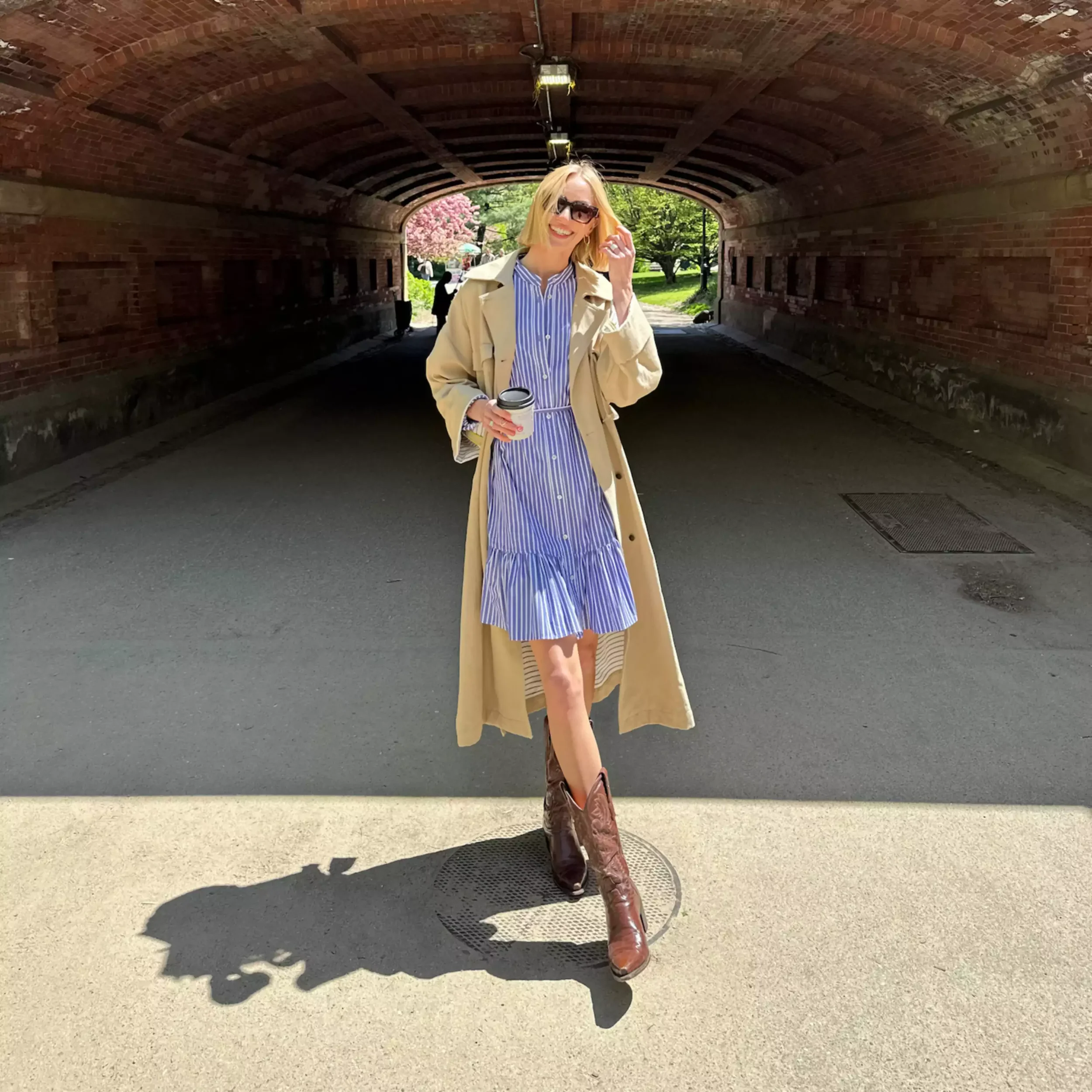 A woman in a blue and white striped dress, beige trench coat, and brown cowboy boots holding a coffee cup, standing under a brick tunnel on a sunny day.
