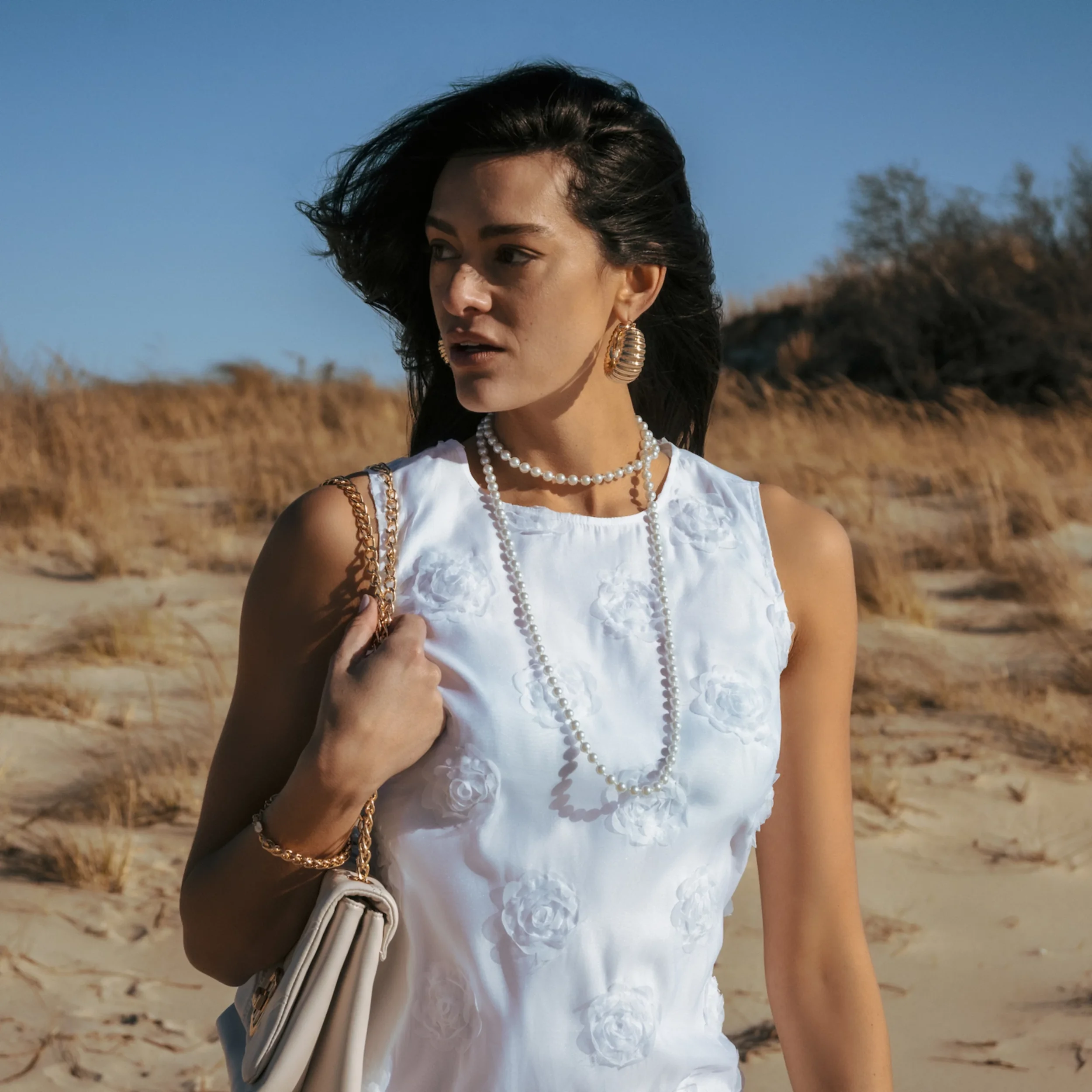 A woman with dark hair and gold jewelry standing on a sandy beach with dry grass and trees in the background.