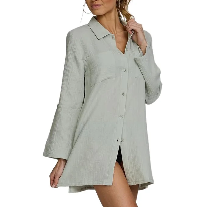 Woman wearing a light gray, button-up shirt dress with long sleeves, standing with her hand near her neck.