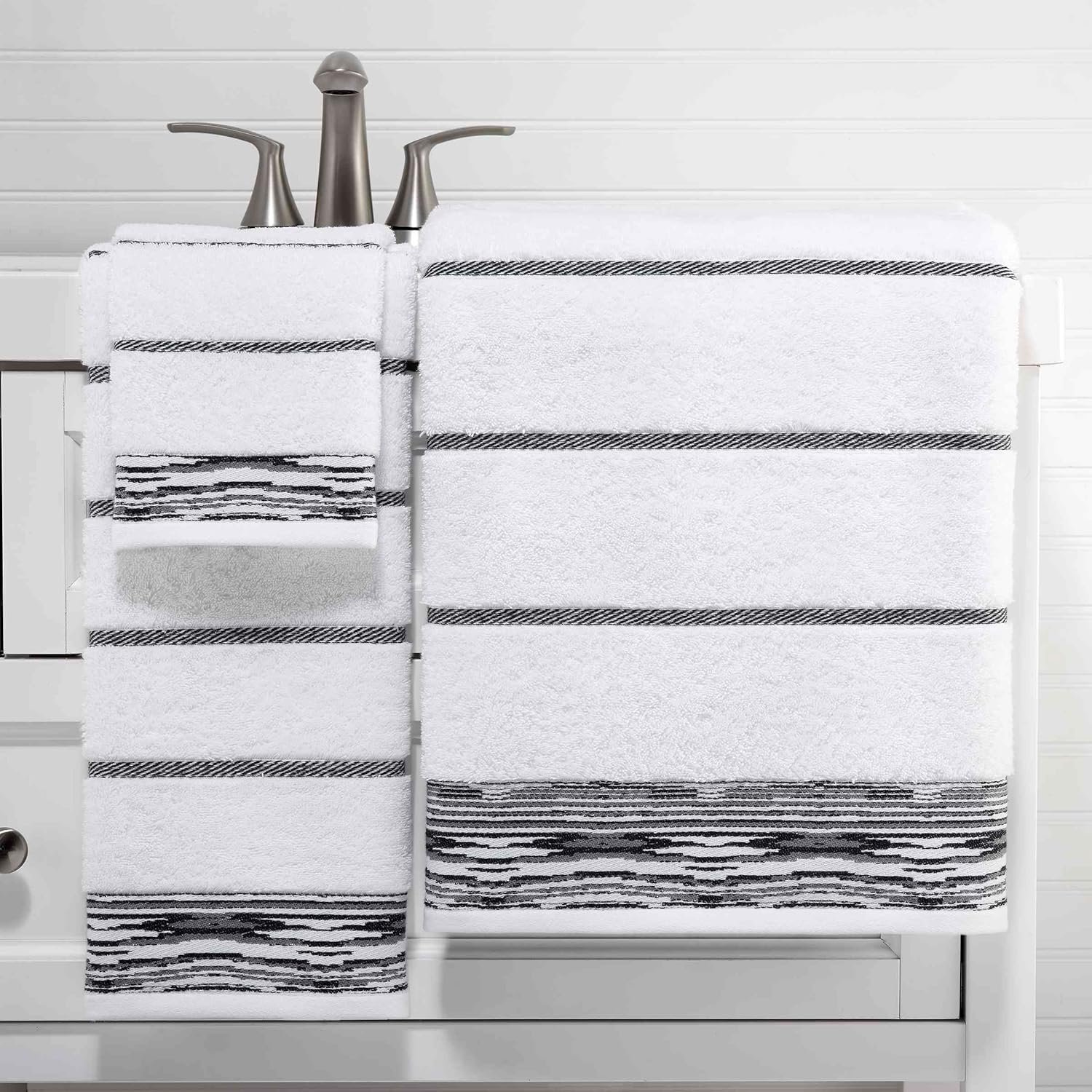 Stacked white towels with black and white striped borders on a white surface in front of a brushed nickel bathroom faucet.