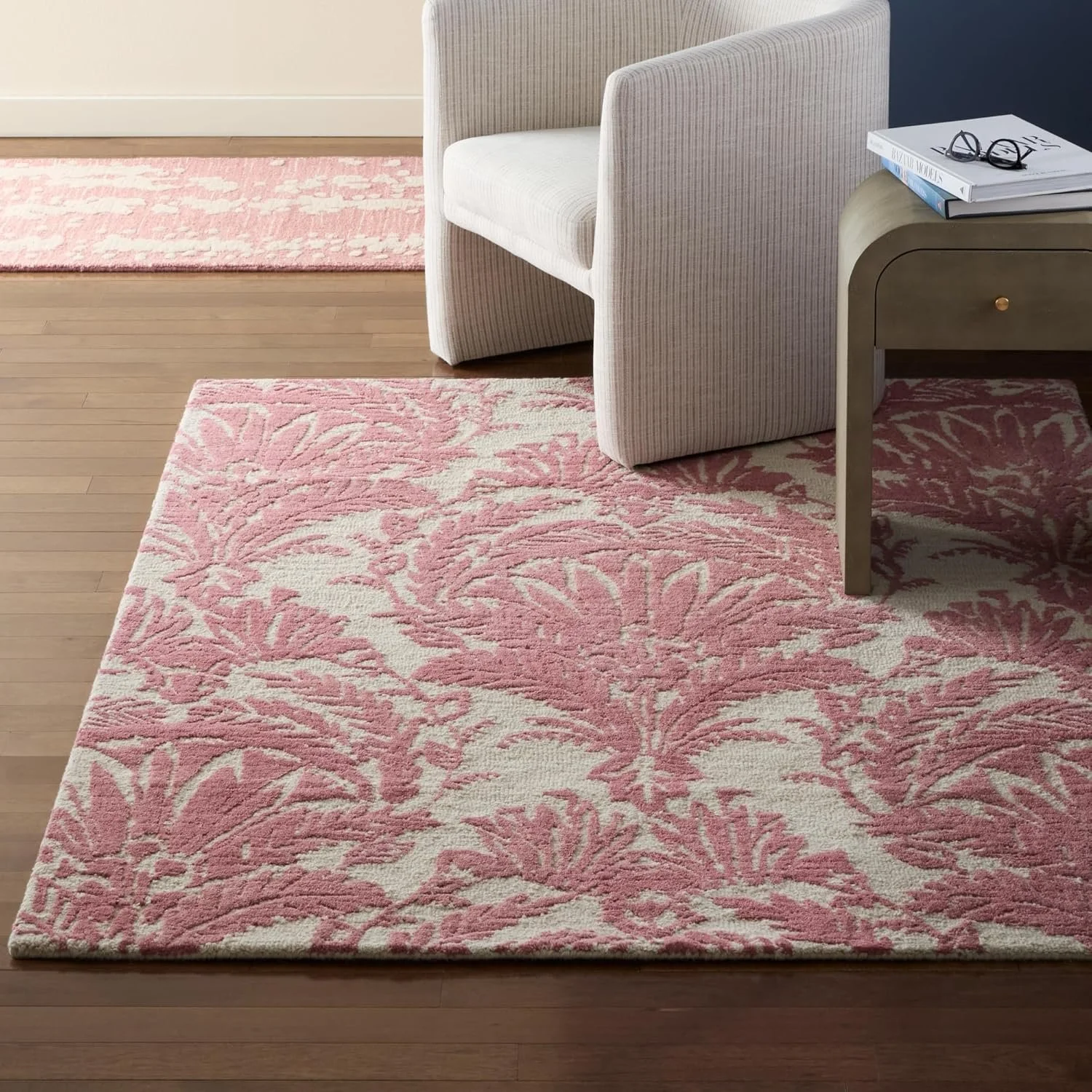 A pink and white floral patterned area rug on a wooden floor next to a light-colored armchair and a side table with books and glasses.