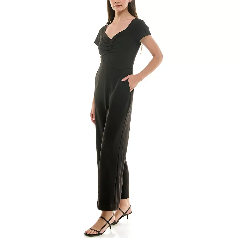 A woman wearing a black short-sleeve jumpsuit with a V-neckline and cinched waist, paired with black strappy high heels.