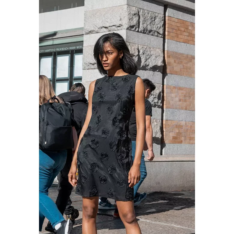 A young woman with shoulder-length black hair in an asymmetrical black dress with floral patterns stands on a city sidewalk near a stone and brick building, with several pedestrians in the background.