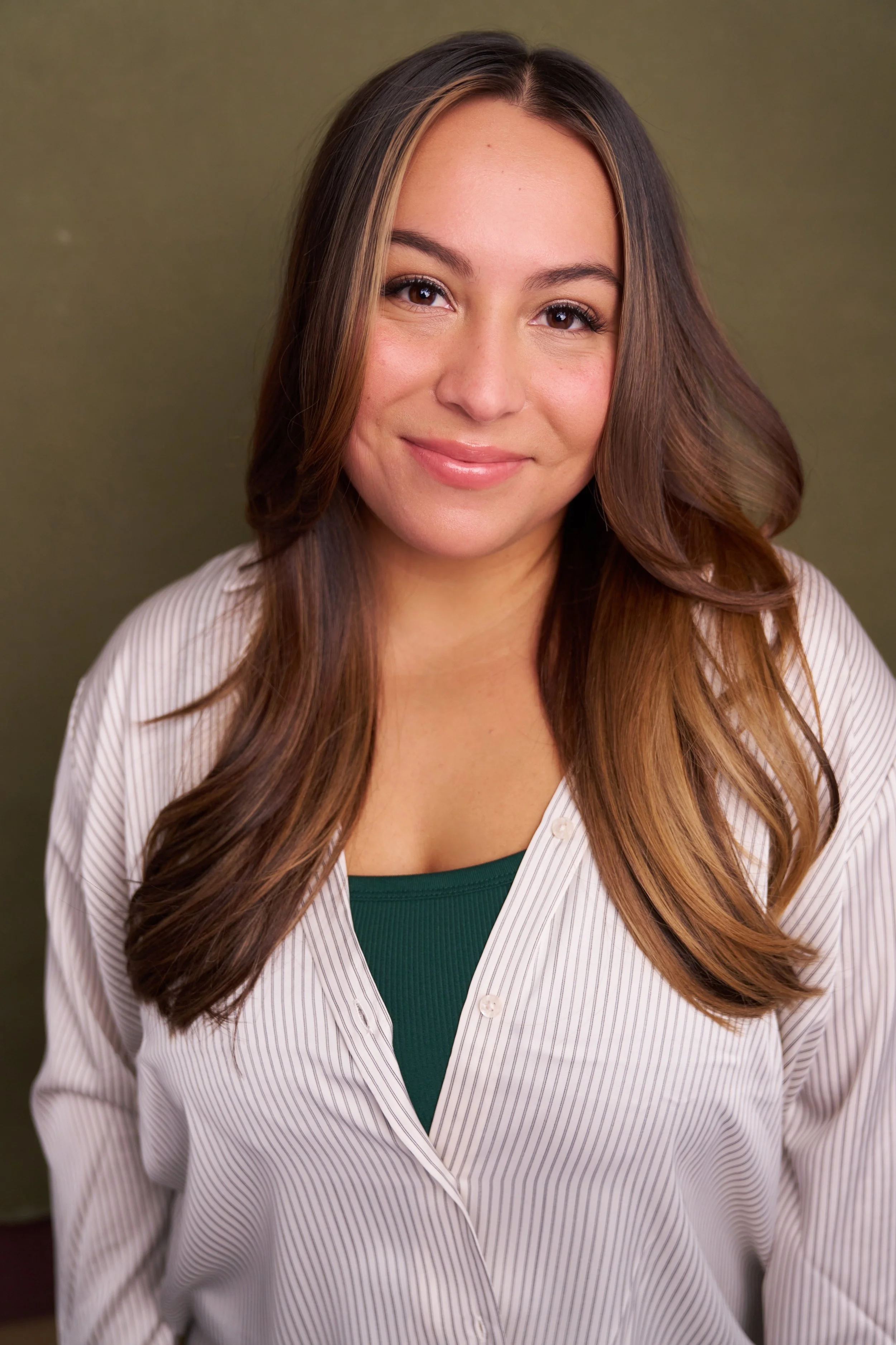 A young woman with long, wavy brown hair wearing a white striped shirt and a green top, smiling at the camera, with a plain olive-green background.