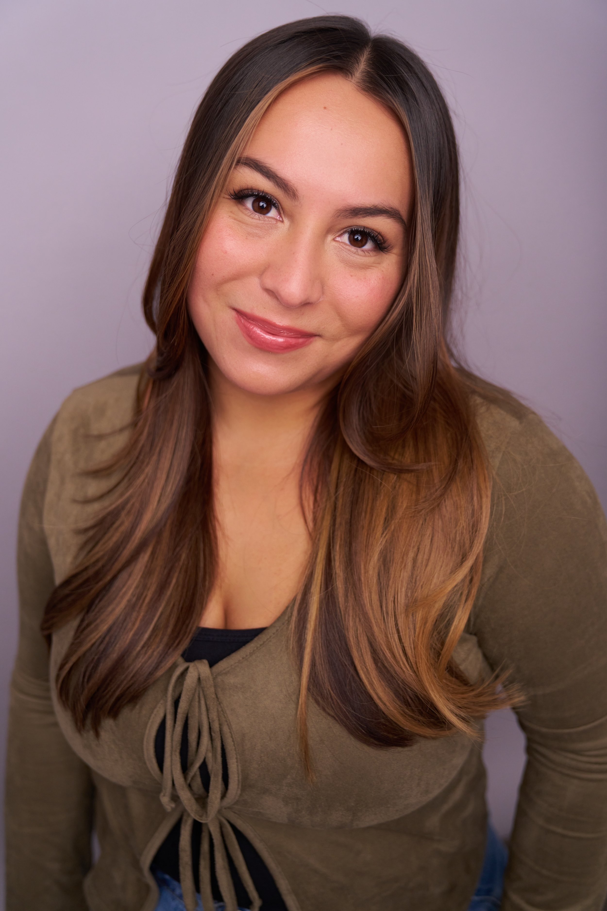 Close-up of a smiling woman with long brown hair, wearing a brown shirt, against a neutral background.