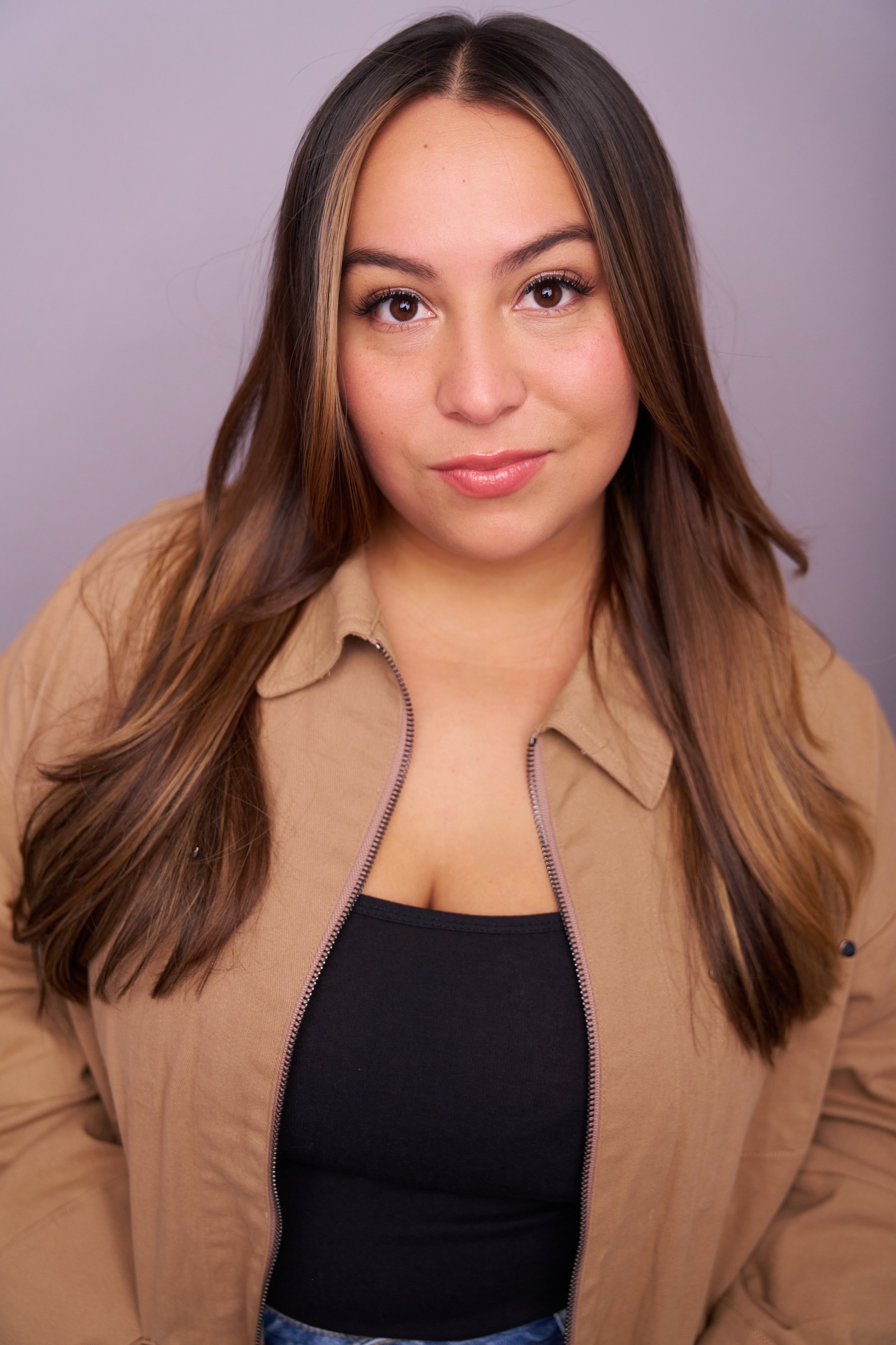 A young woman with long brown hair, wearing a black top and tan jacket, looking directly at the camera with a slight smile.