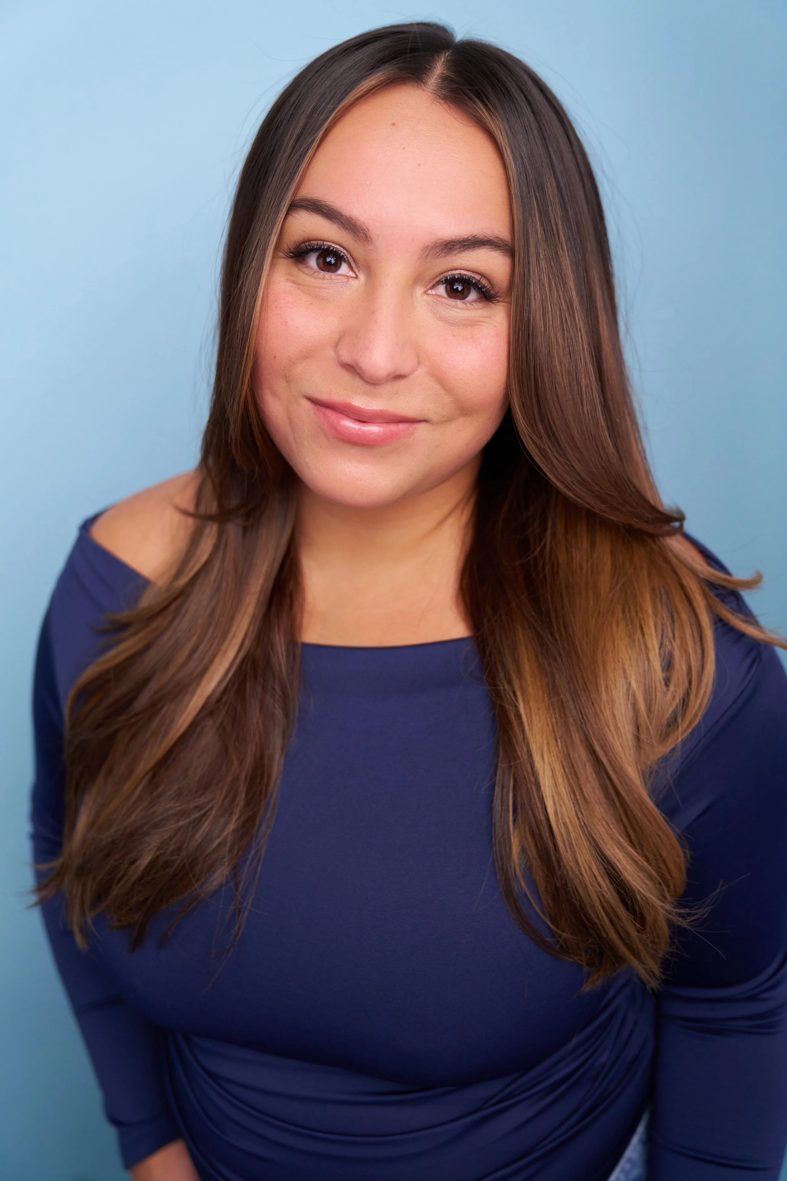 Portrait of a woman with long brown hair, wearing a navy blue top, smiling against a light blue background.