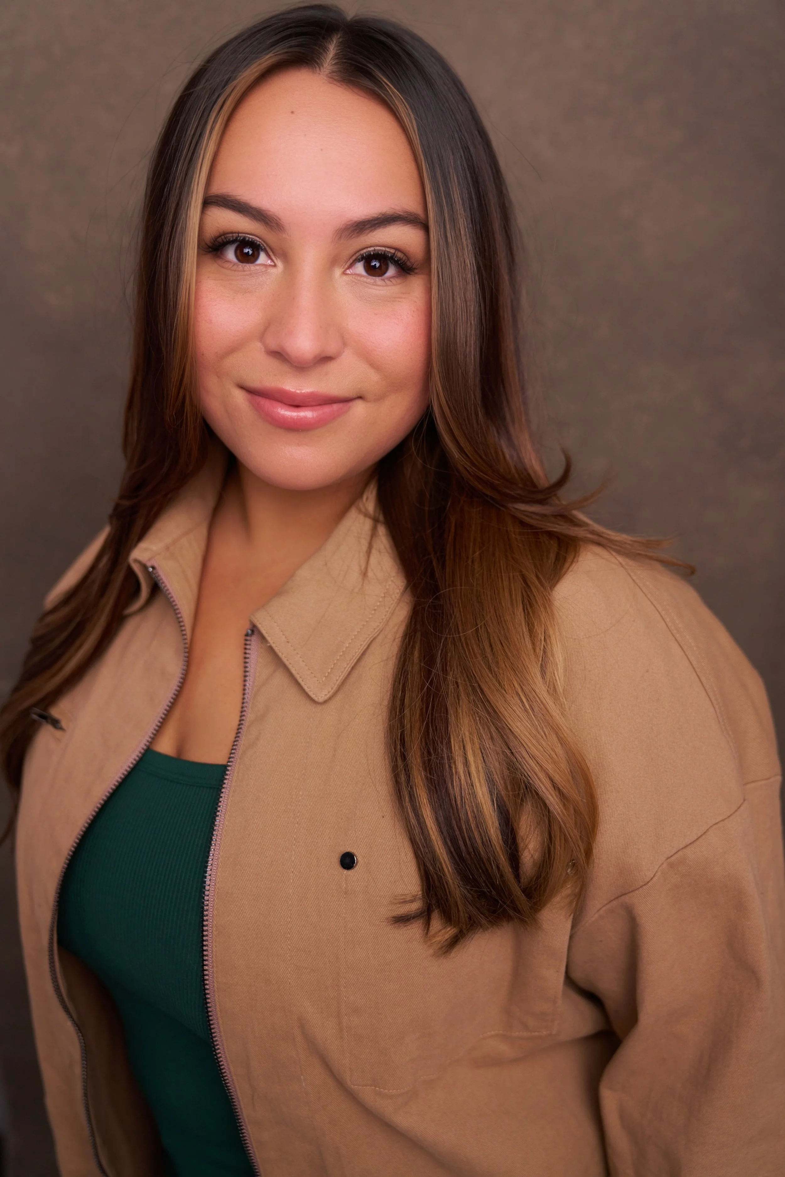 A woman with long brown hair, wearing a beige jacket over a green top, looking at the camera with a slight smile, against a neutral background.