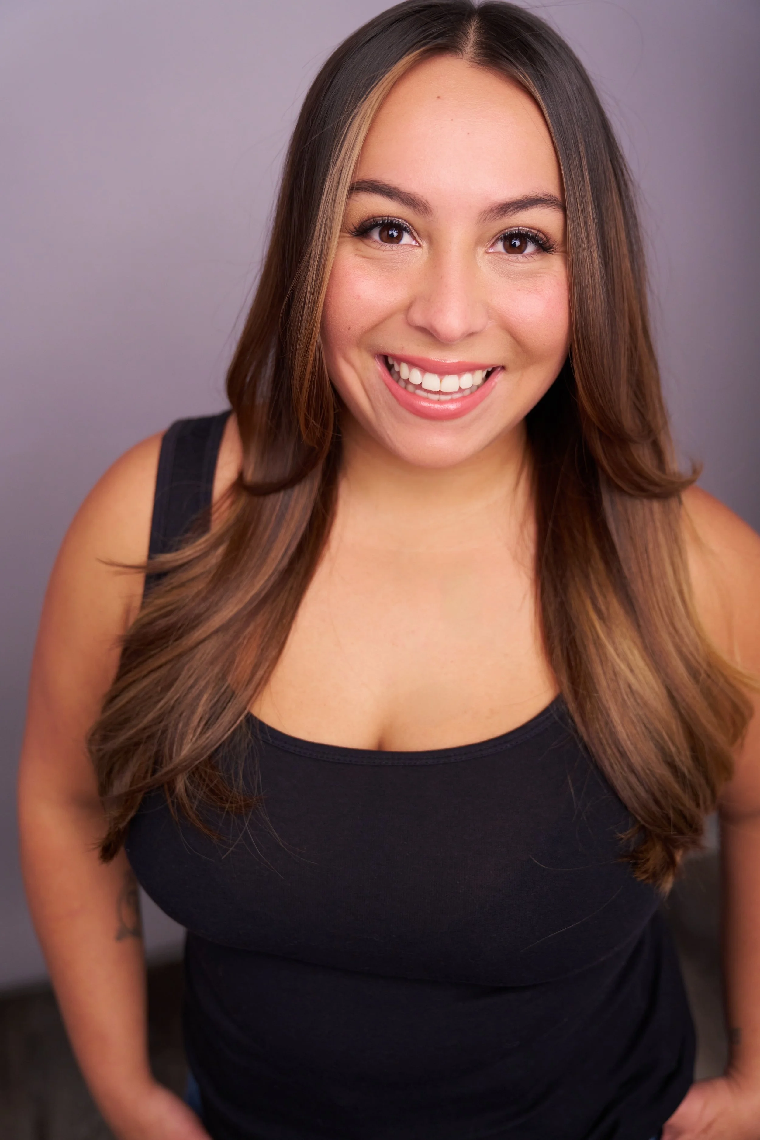 A smiling young woman with long wavy brown hair, wearing a black sleeveless top, looking directly at the camera against a plain neutral background.