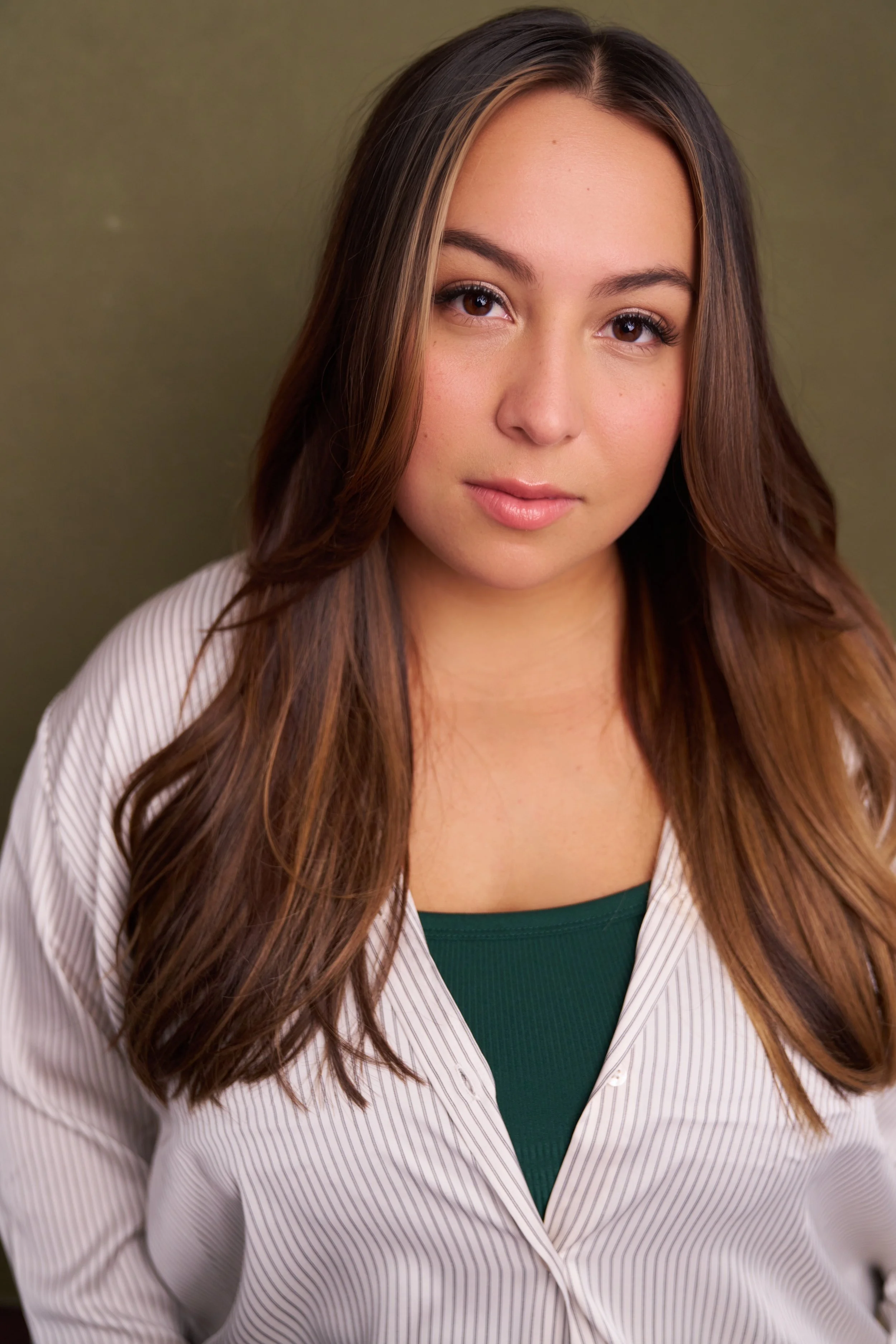 A young woman with long brown hair and light skin looking at the camera, wearing a white and light pink striped shirt over a green top, with a neutral background.