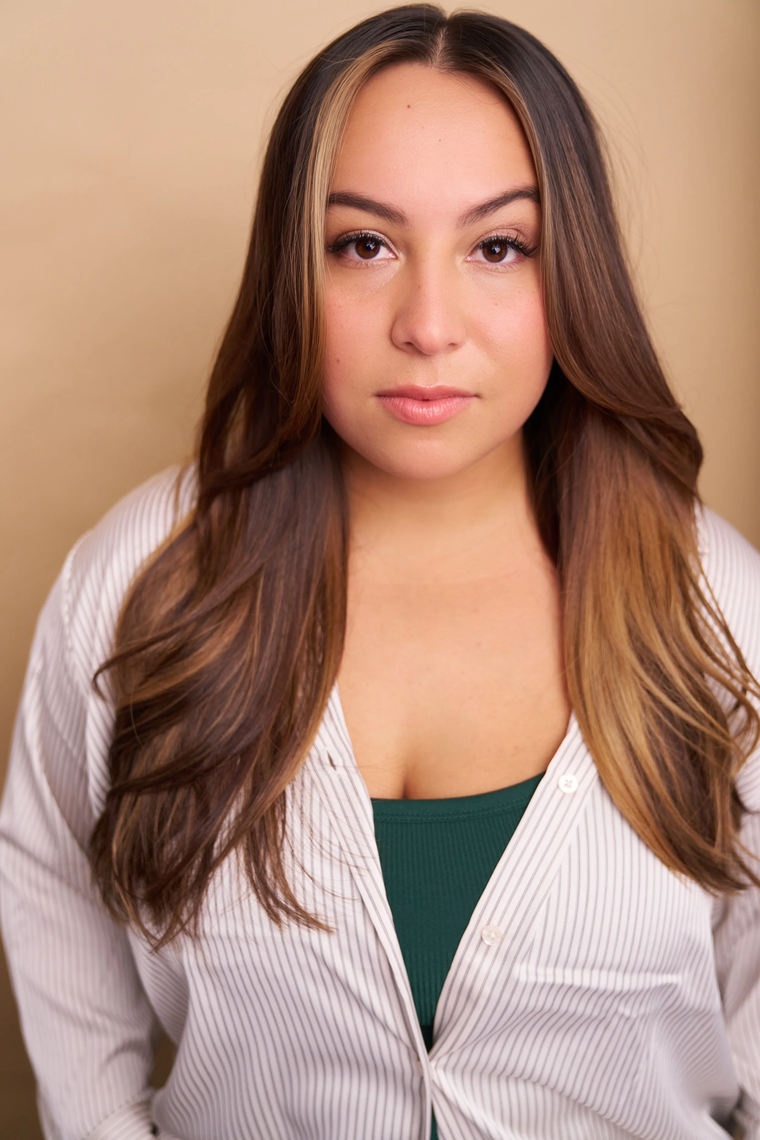 Close-up portrait of a young woman with long brown hair and light skin, wearing a green top and a white striped shirt, standing against a beige background.