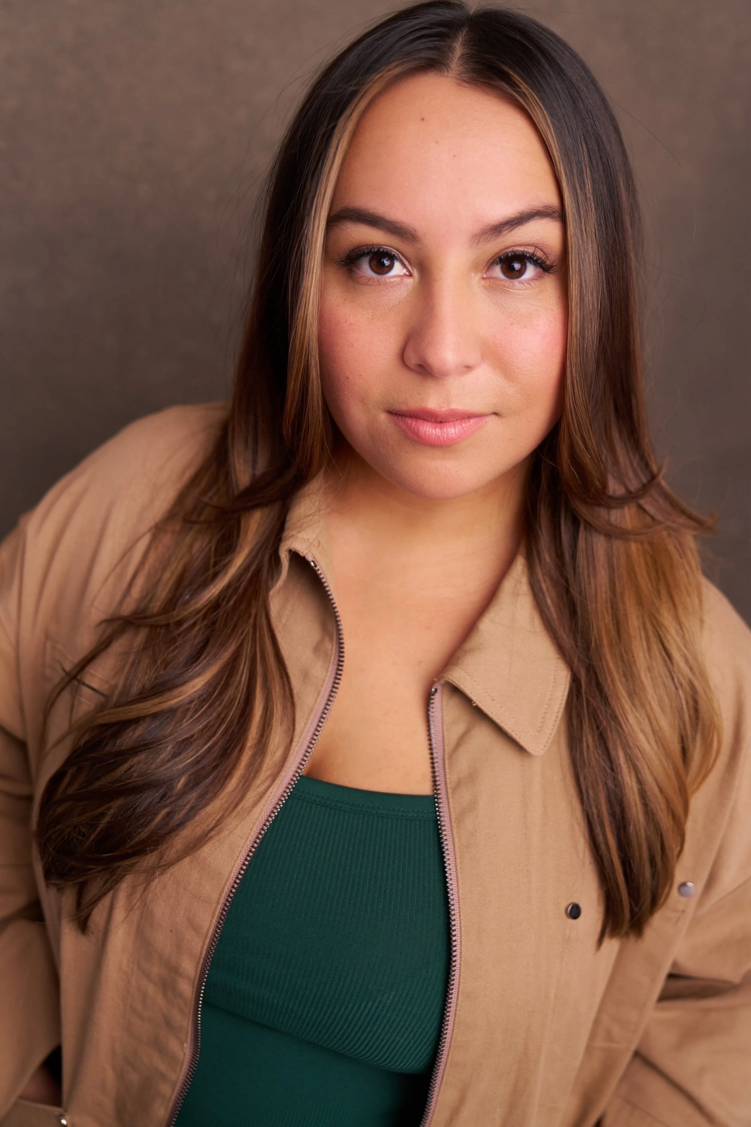 Close-up portrait of a young woman with long brown hair, wearing a beige jacket over a green top, against a plain background.