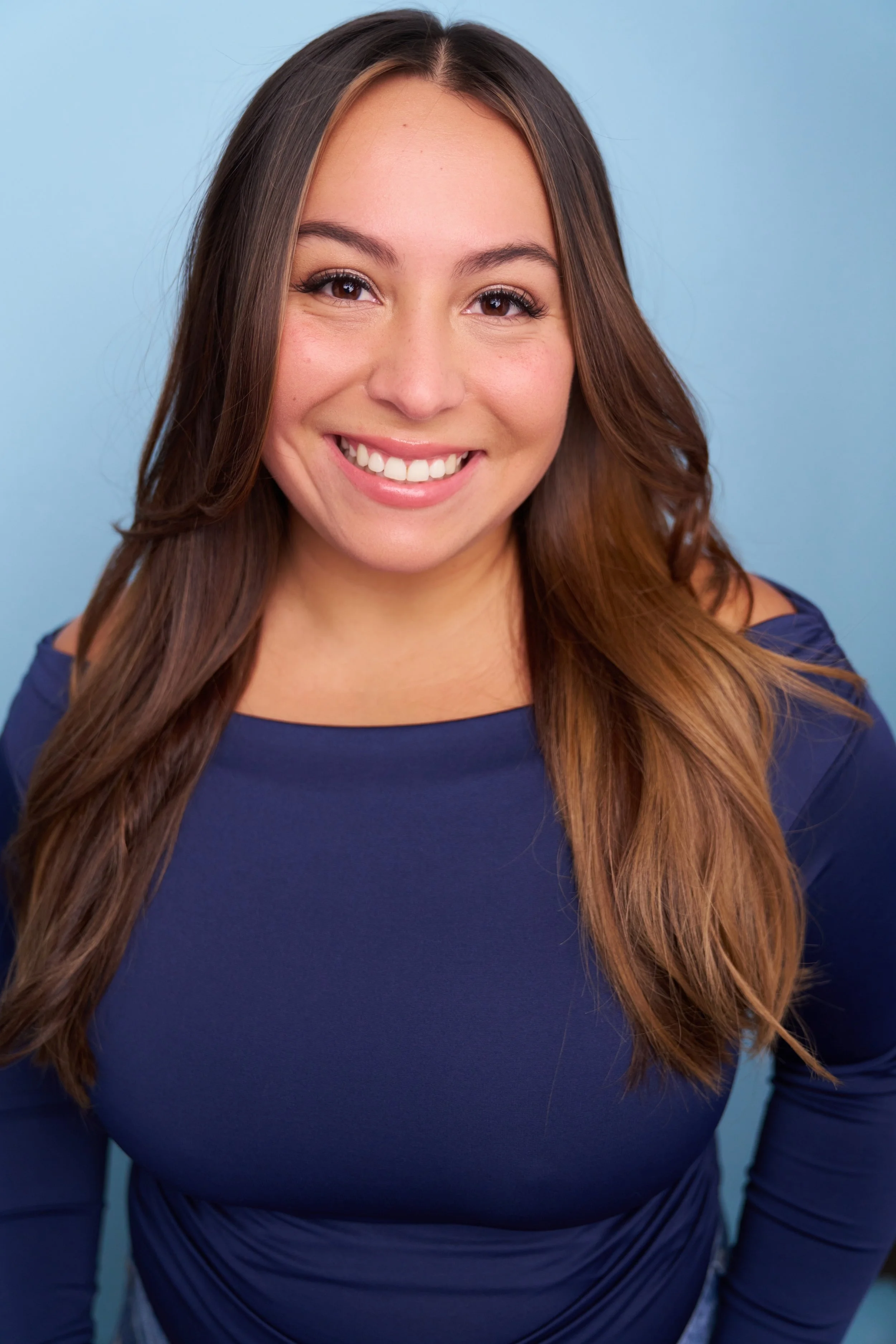 Portrait of a young woman with long brown hair wearing a blue top, smiling against a light blue background.