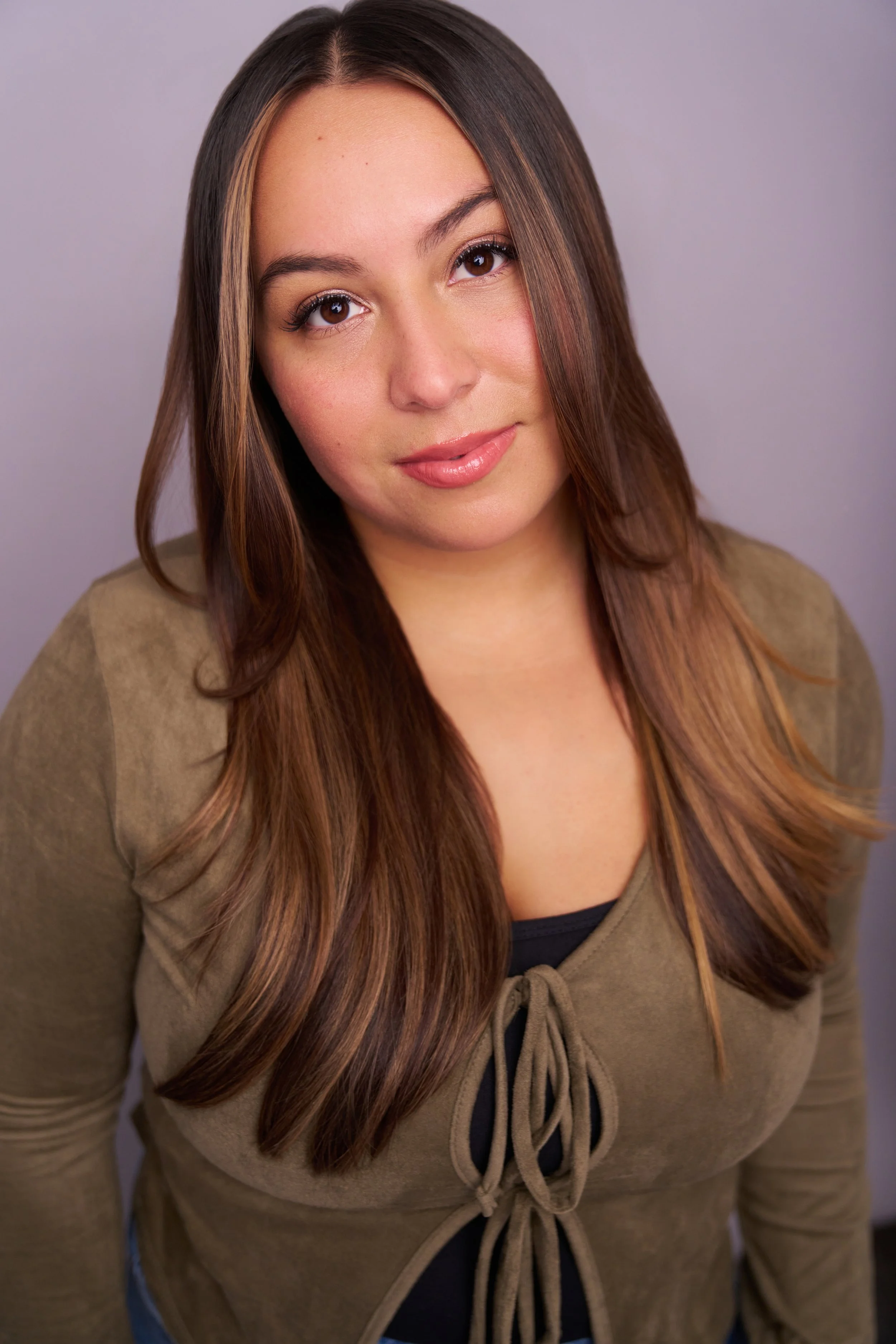 A young woman with long brown hair and a slight smile, wearing a tan jacket over a black top, posing against a plain gray background.