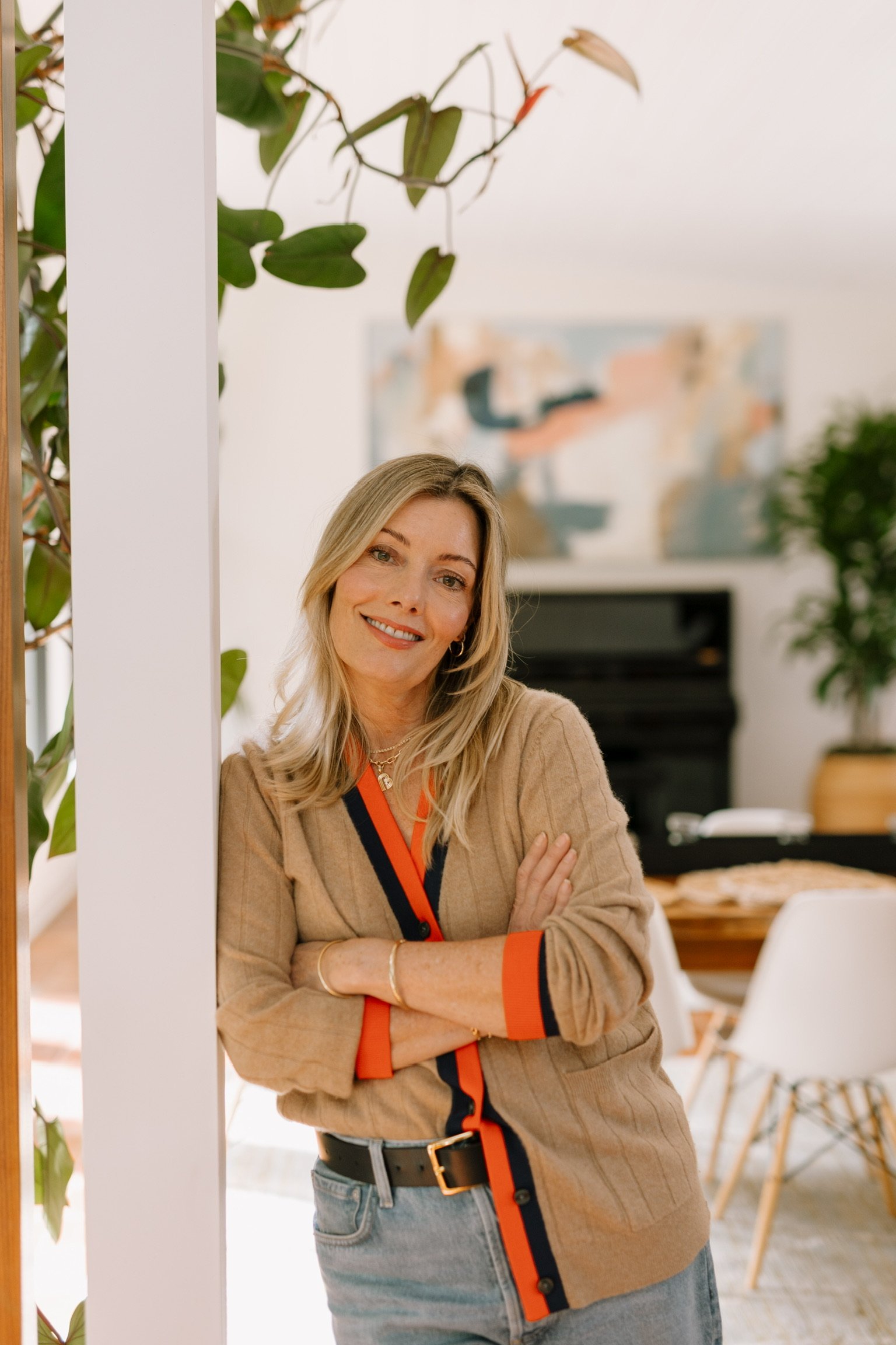 Betsy leaning against the wall smiling at the camera in a naturally lit living room with plants and paintings around her