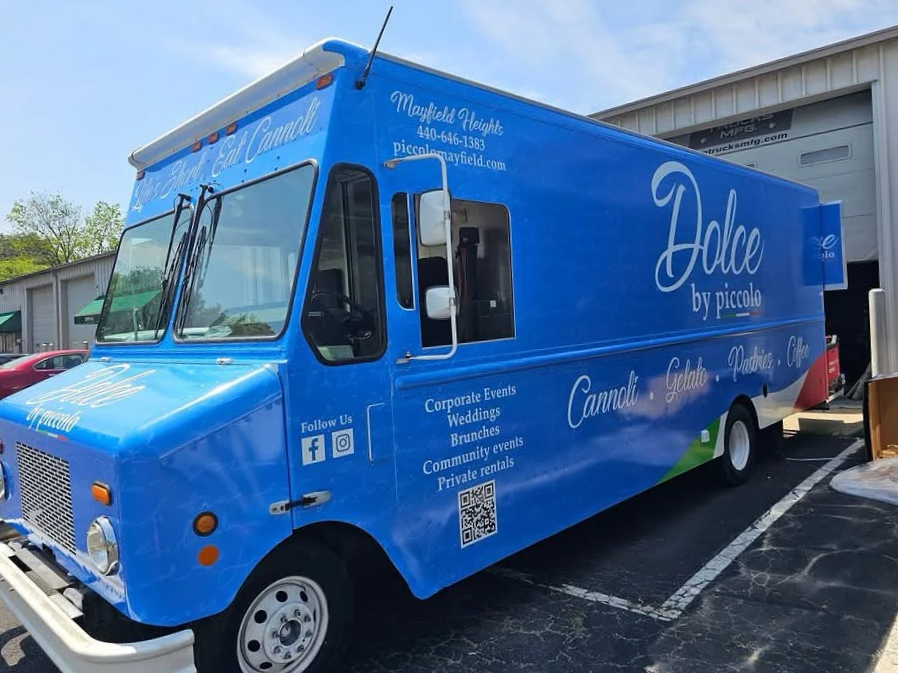 A blue food truck with the words "Dolce by piccolo" on its side, parked in front of a building. The truck lists services like corporate events, weddings, brunches, community events, and private rentals, with social media icons and a QR code.