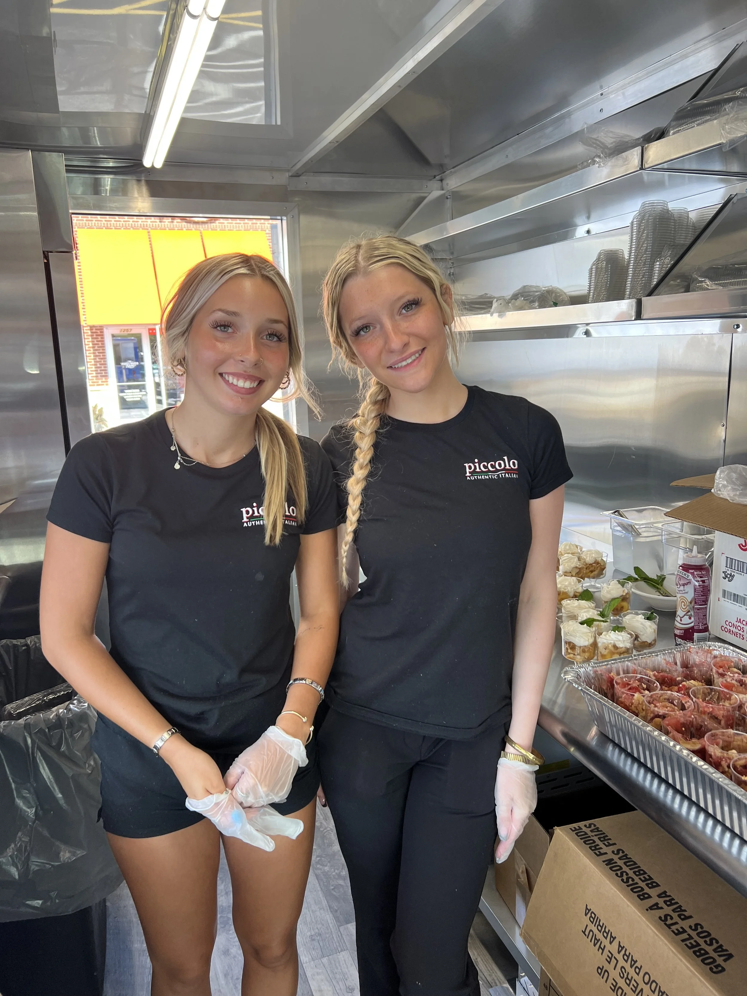Two young women in black t-shirts with 'piccolo' logo, smiling and wearing gloves, standing in a food stand with containers of strawberries and desserts around them.