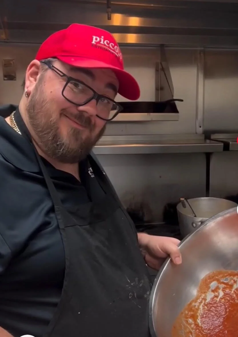 A man wearing glasses, a red cap, and a black apron holds a large metal bowl containing sauce, standing in a commercial kitchen.
